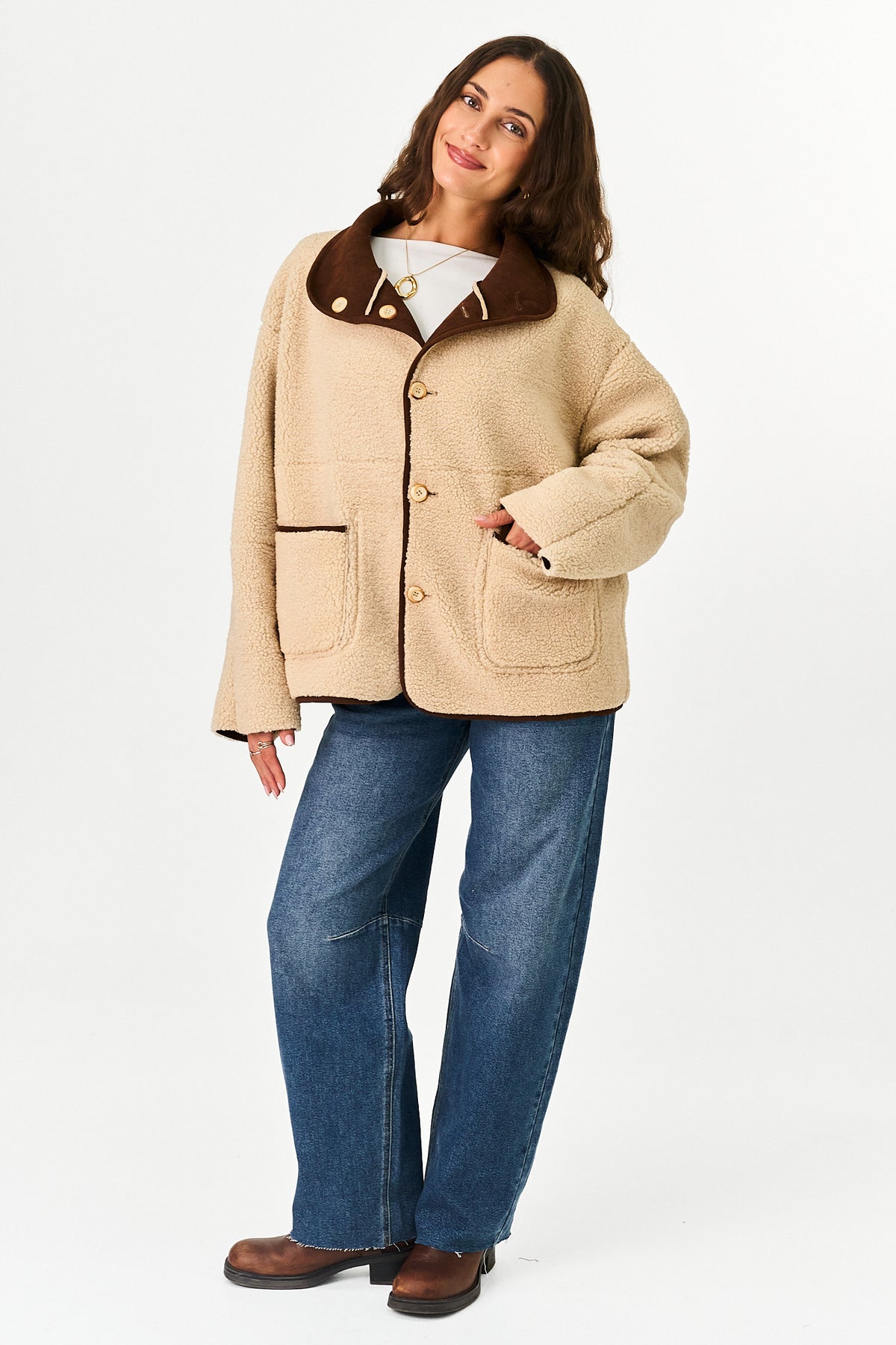 A person wearing a light brown sherpa-like jacket with a brown suede collar and contrast beige piping, paired with blue jeans, against a plain white studio background.