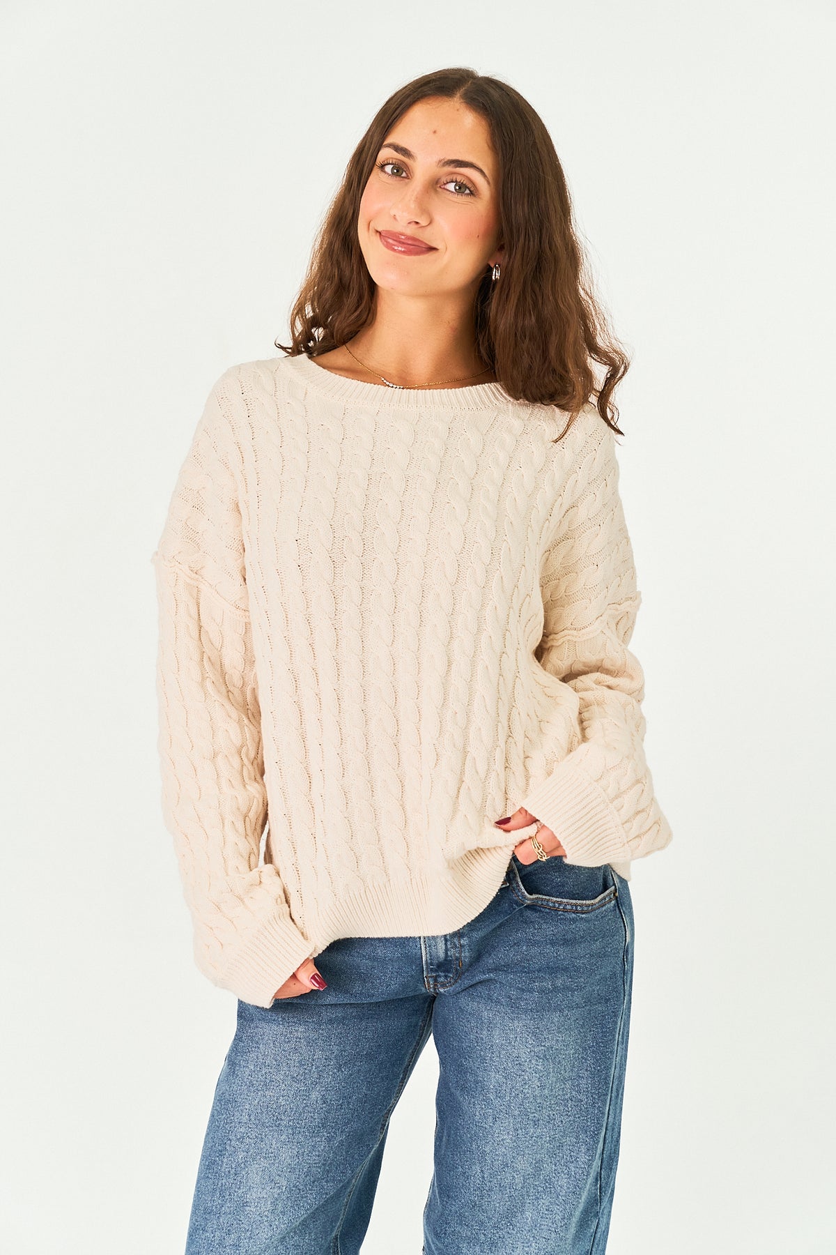 A woman wearing a cream-colored cable-knit sweater and blue jeans, standing against a plain white studio background.