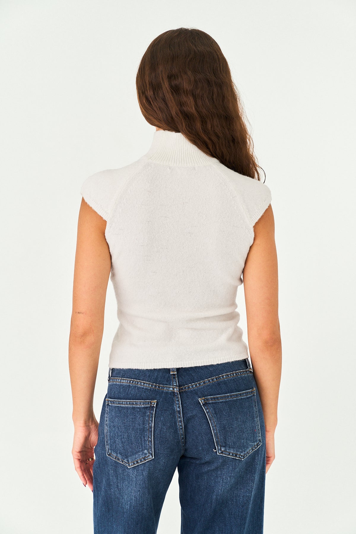 A woman viewed from the back wearing a fitted off-white ribbed knit mock-neck top with cap sleeves and blue jeans, against a plain white studio background.