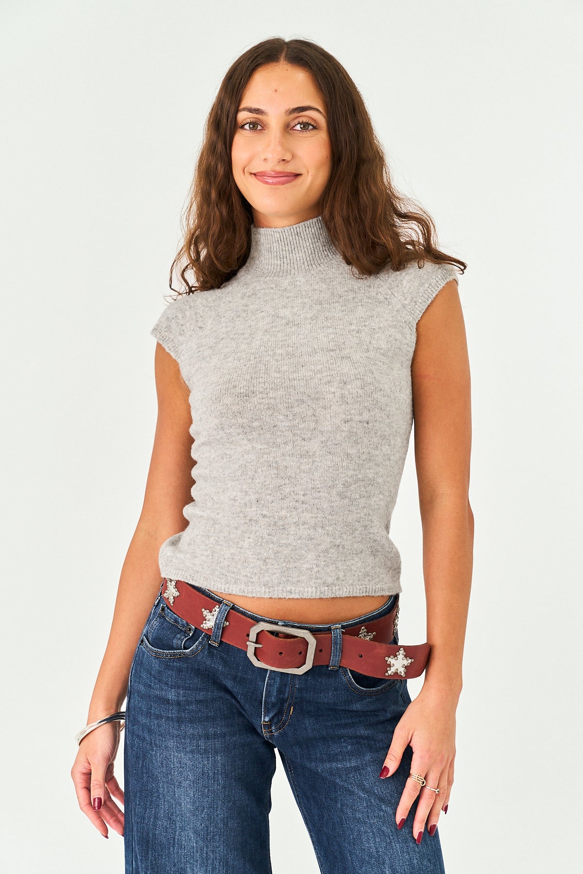 A woman modeling a light gray ribbed knit mock neck top with cap sleeves, paired with blue jeans and a brown western-style belt with silver star details, against a plain white studio background.