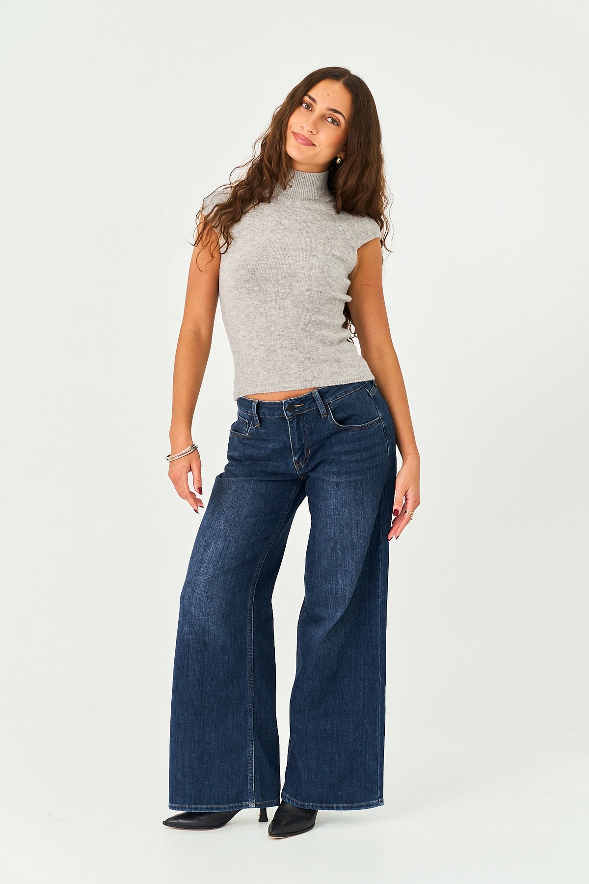 A woman modeling a pair of dark blue wide-leg jeans with a gray sleeveless mock-neck top, standing against a plain white studio background.