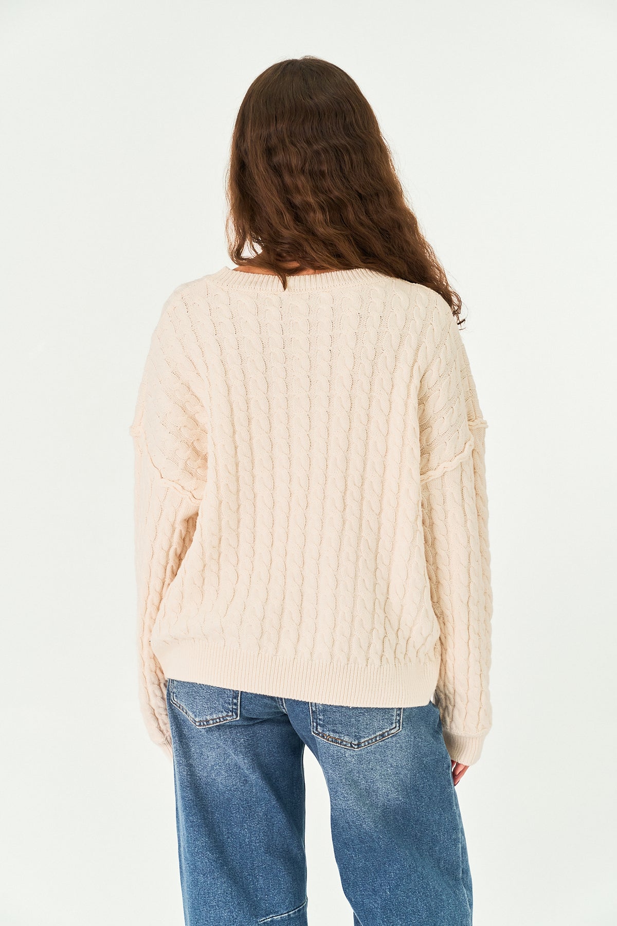 A woman viewed from behind wearing a cream-colored cable-knit sweater and blue jeans against a plain white background.