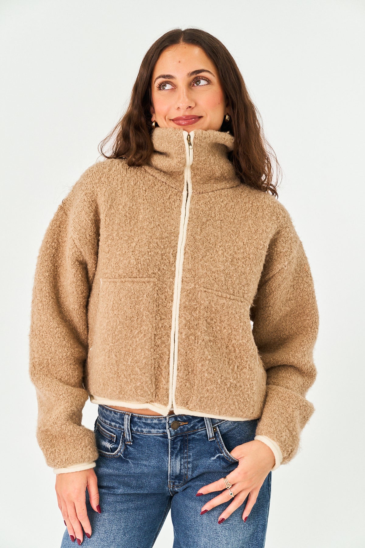 A woman wearing a cropped, high-collar teddy fleece jacket in a warm camel beige color with a cream zipper and cuffs, paired with blue jeans, against a plain white studio background.