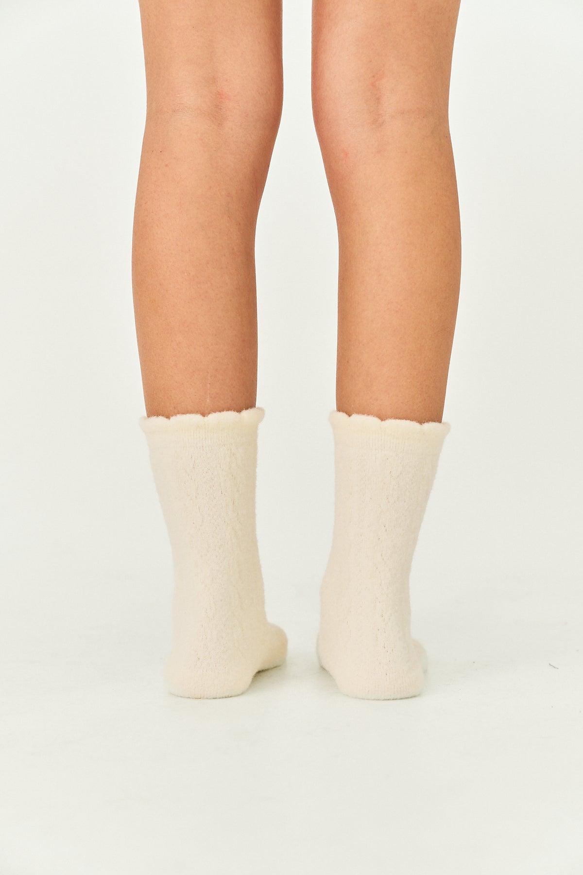 A pair of cream-colored, fuzzy crew socks with scalloped edges worn on a person's legs, shown from the back against a plain white studio background.