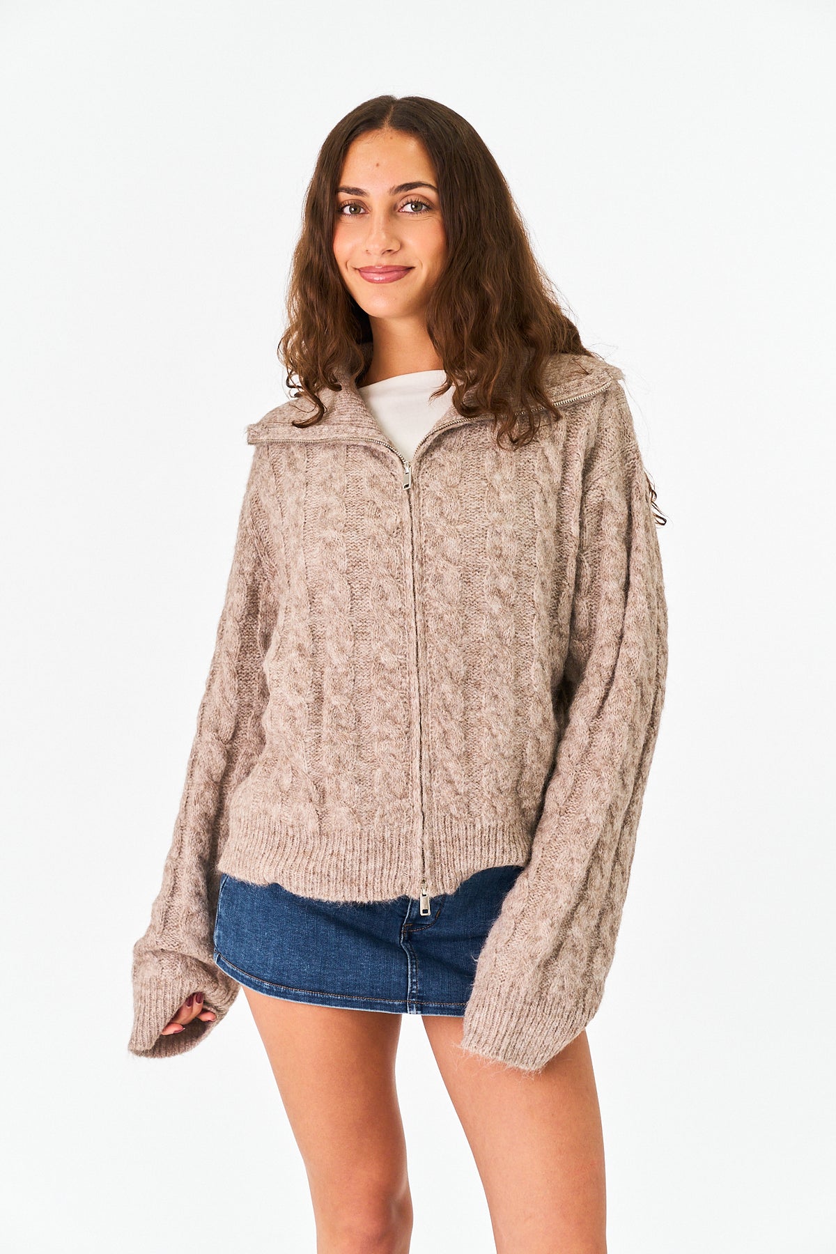 A woman modeling a chunky cable-knit zip-up hoodie in a heathered taupe-beige color, styled with a white t-shirt and a blue denim mini skirt against a plain white studio background.