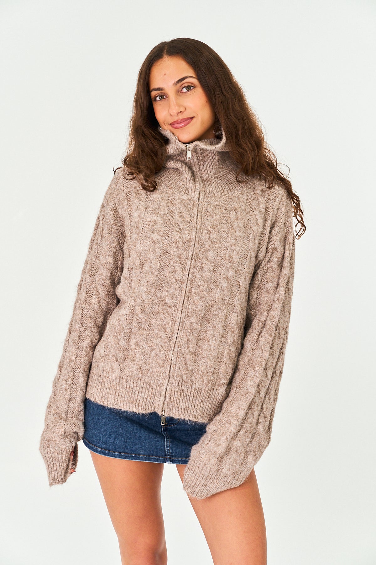 A woman modeling a chunky cable-knit zip-up sweater in a heathered taupe-beige color, paired with a blue denim mini skirt, against a plain white studio background.