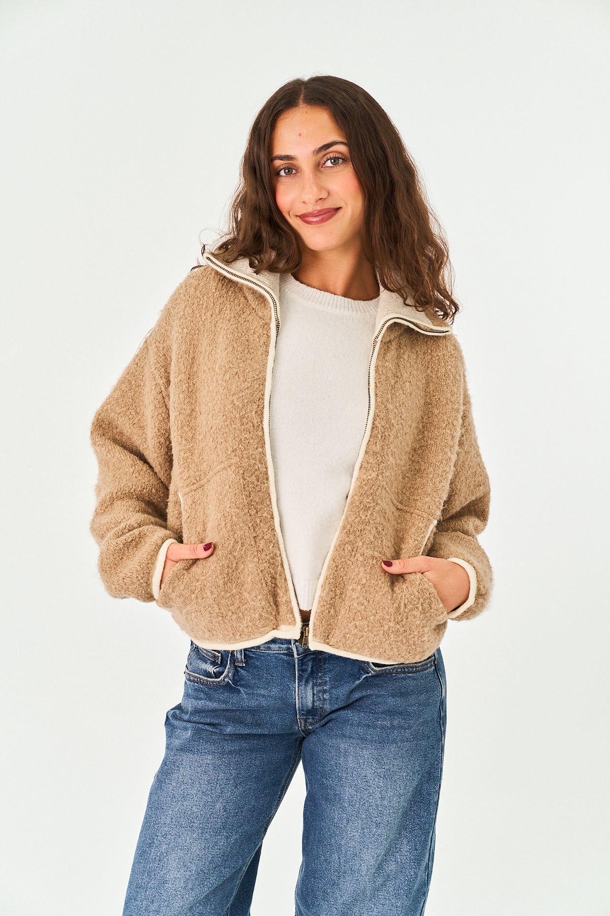 A woman modeling a beige sherpa fleece zip-up jacket with cream trim, worn over a white top and blue jeans, against a plain white studio background.