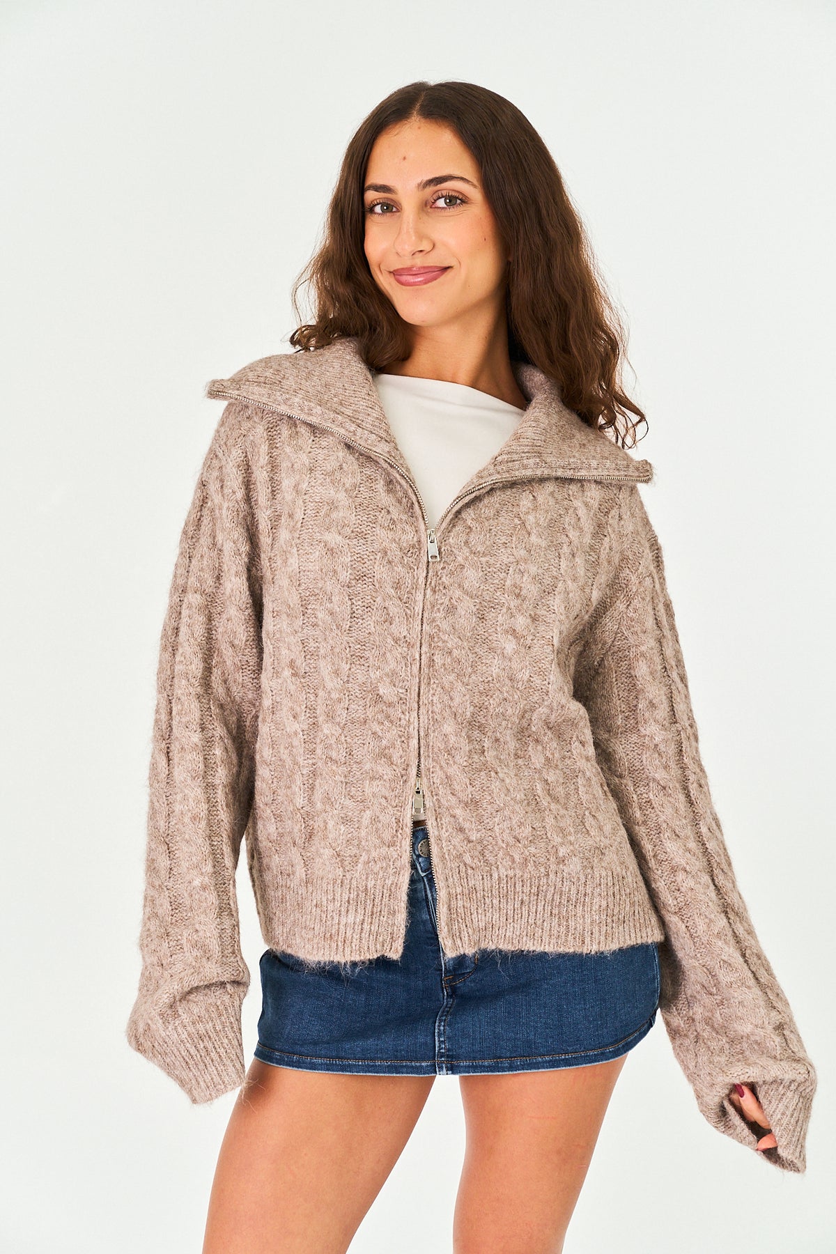 A woman modeling a chunky knit zip-up cardigan in a heathered taupe-beige color, styled with a white top and a blue denim mini skirt against a plain white studio background.