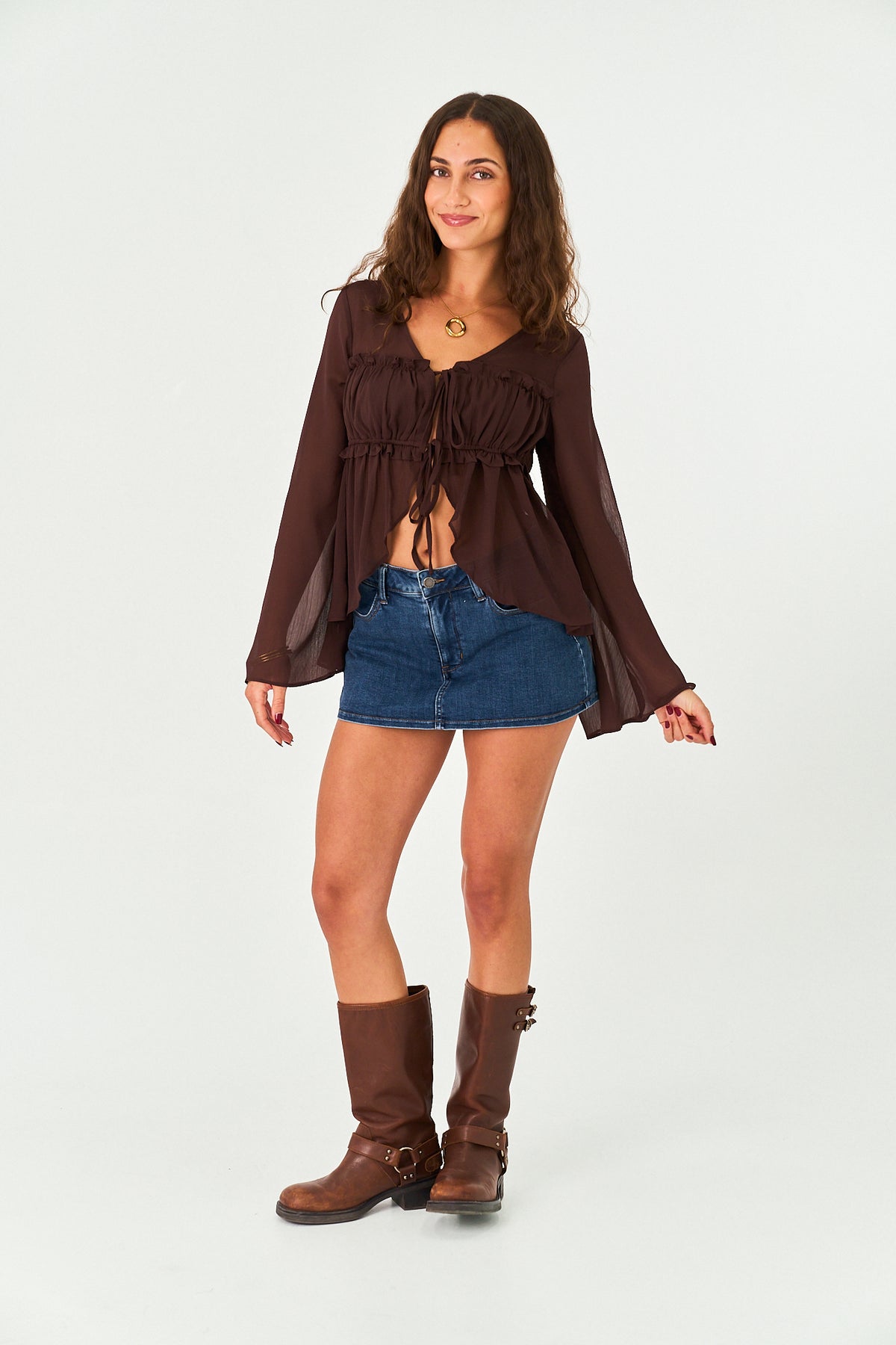 A woman modeling a dark brown sheer long-sleeve crop top with a tie-front detail, paired with a denim mini skirt and brown boots, against a plain light gray background.
