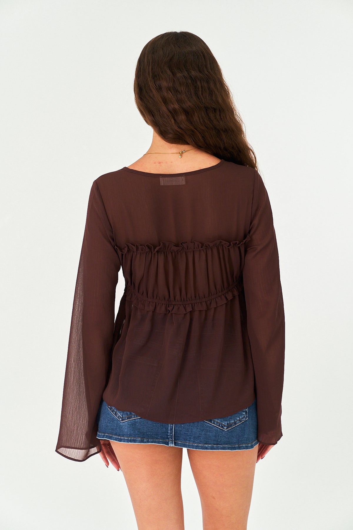 A woman modeling a dark brown sheer long-sleeve tie-front top with a ruffled neckline, paired with a denim mini skirt, against a plain white studio background.