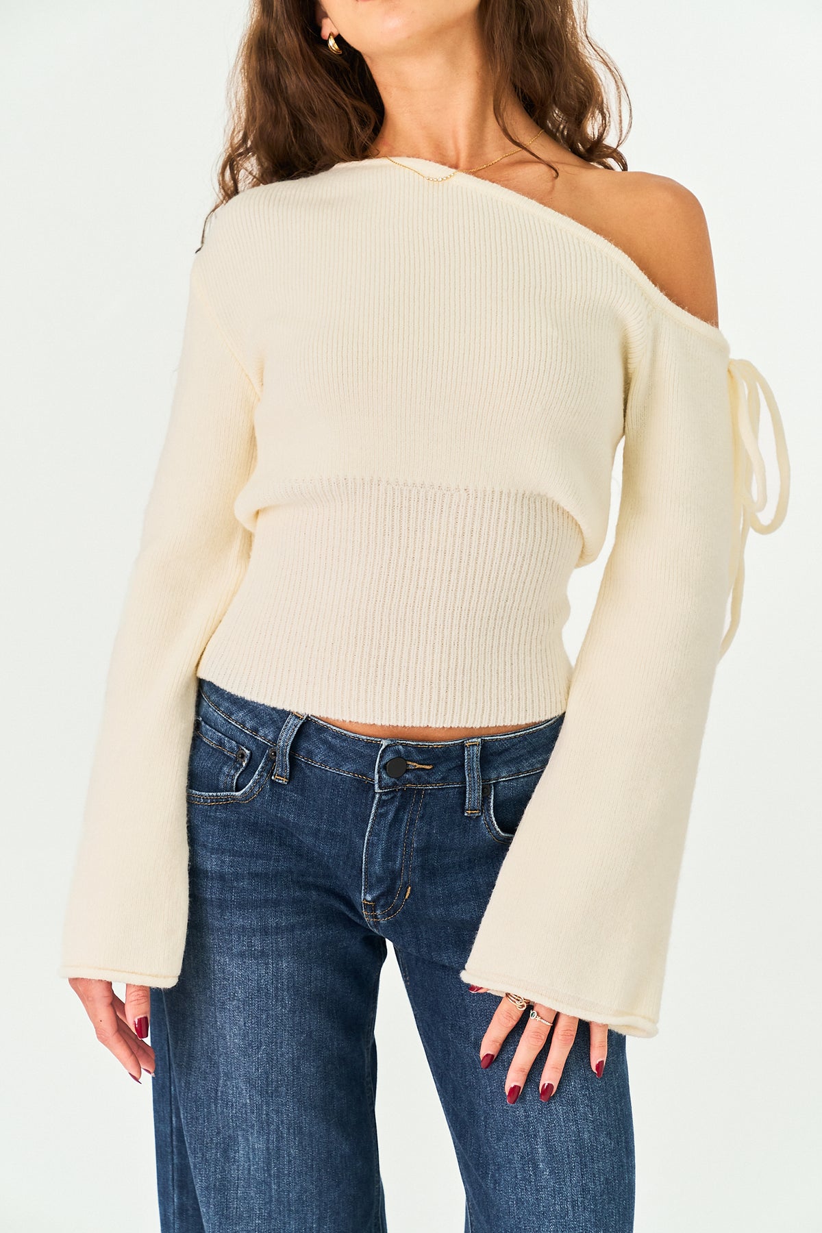 A woman wearing a cream off-the-shoulder ribbed knit sweater with long bell sleeves and side ties, paired with blue jeans, against a plain white background.