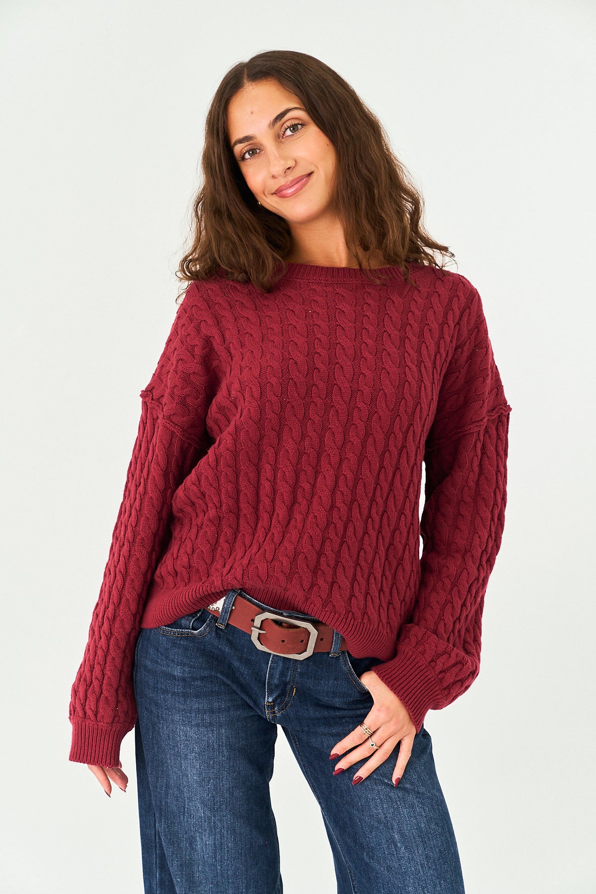 A woman wearing a chunky cable-knit sweater in a deep burgundy red, paired with blue jeans and a brown belt, posing against a light gray studio background.