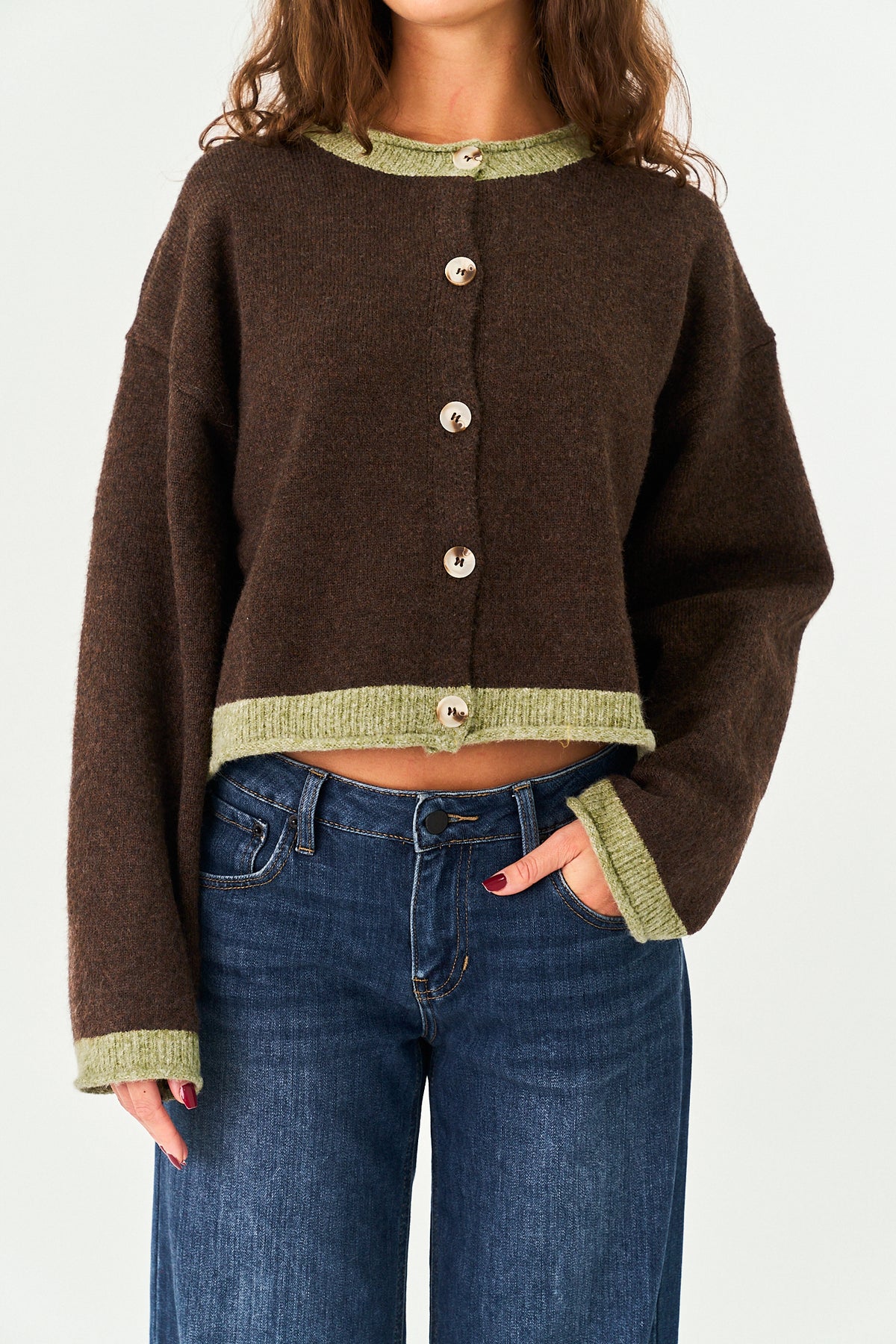 A woman wearing a cropped brown knit cardigan with sage green ribbed trim and buttons, paired with blue jeans, against a plain light background.
