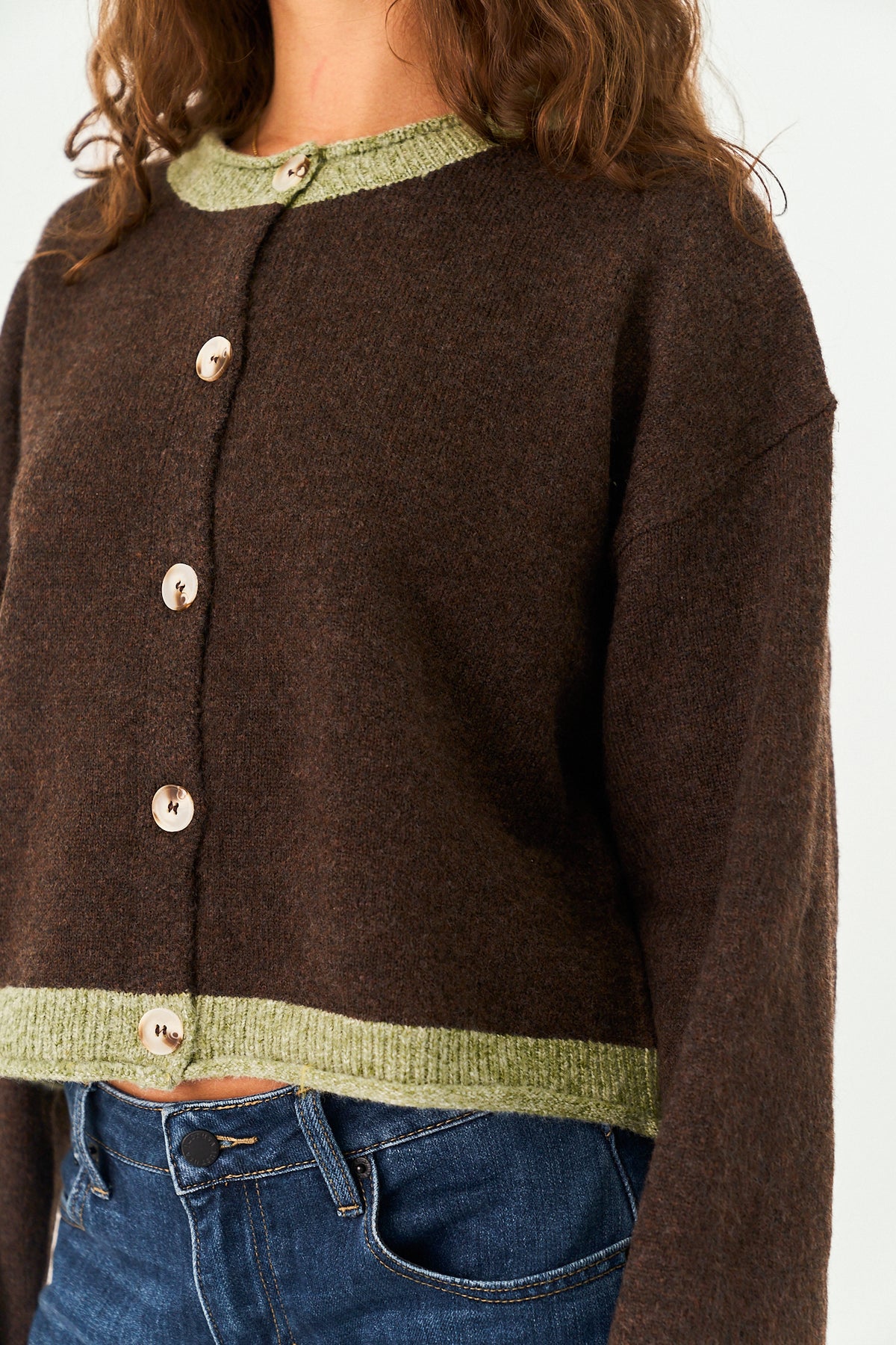 A cropped brown knit cardigan with light olive green ribbed trim at the crew neck and hem, featuring four light-colored buttons, shown on a model wearing blue jeans.