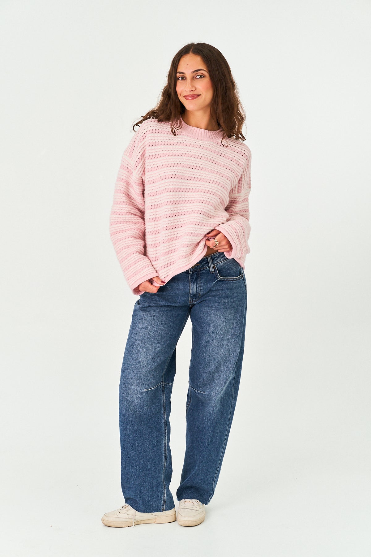A woman modeling a light pink knit sweater with horizontal textured stripes, paired with blue jeans and off-white sneakers, standing against a plain white studio background.