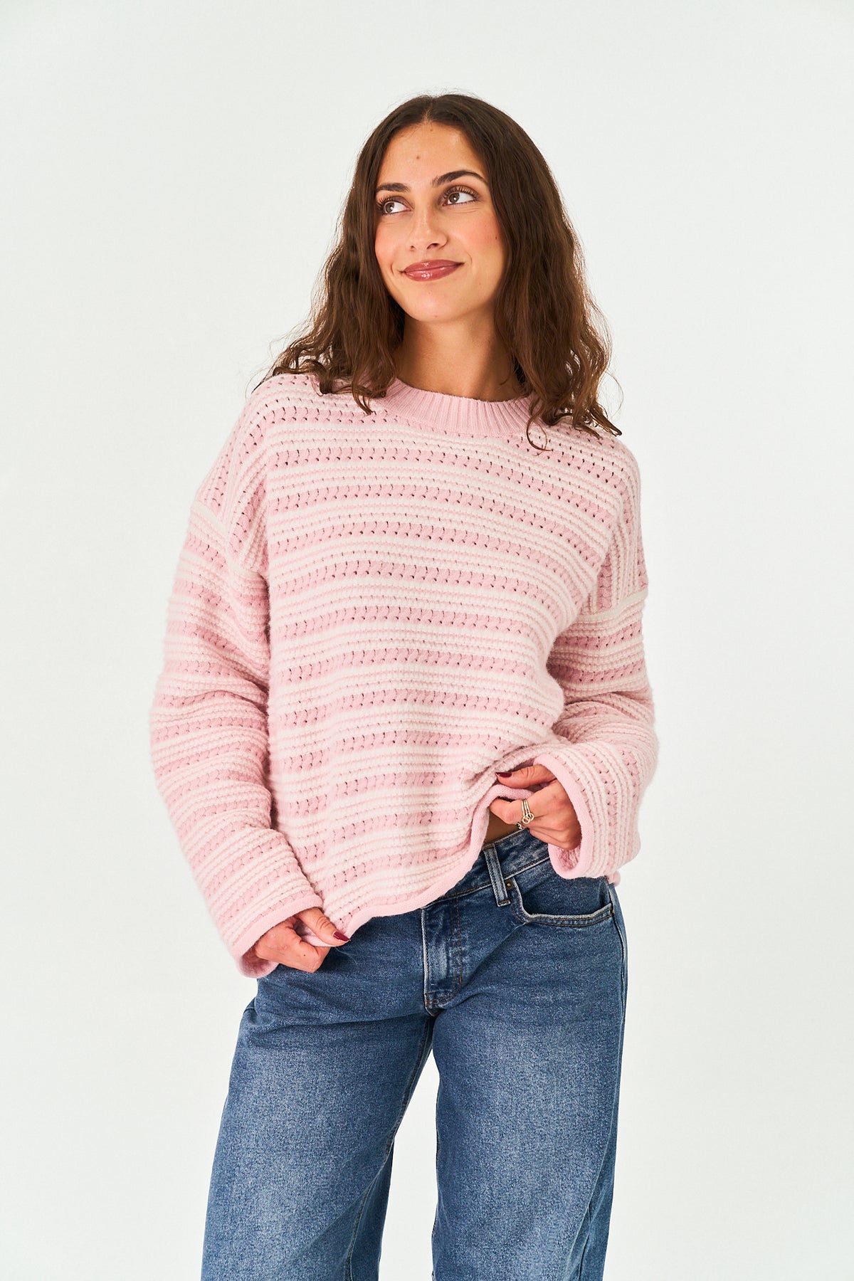 A woman wearing a pink and white striped knit sweater and blue jeans, standing against a plain white studio background.