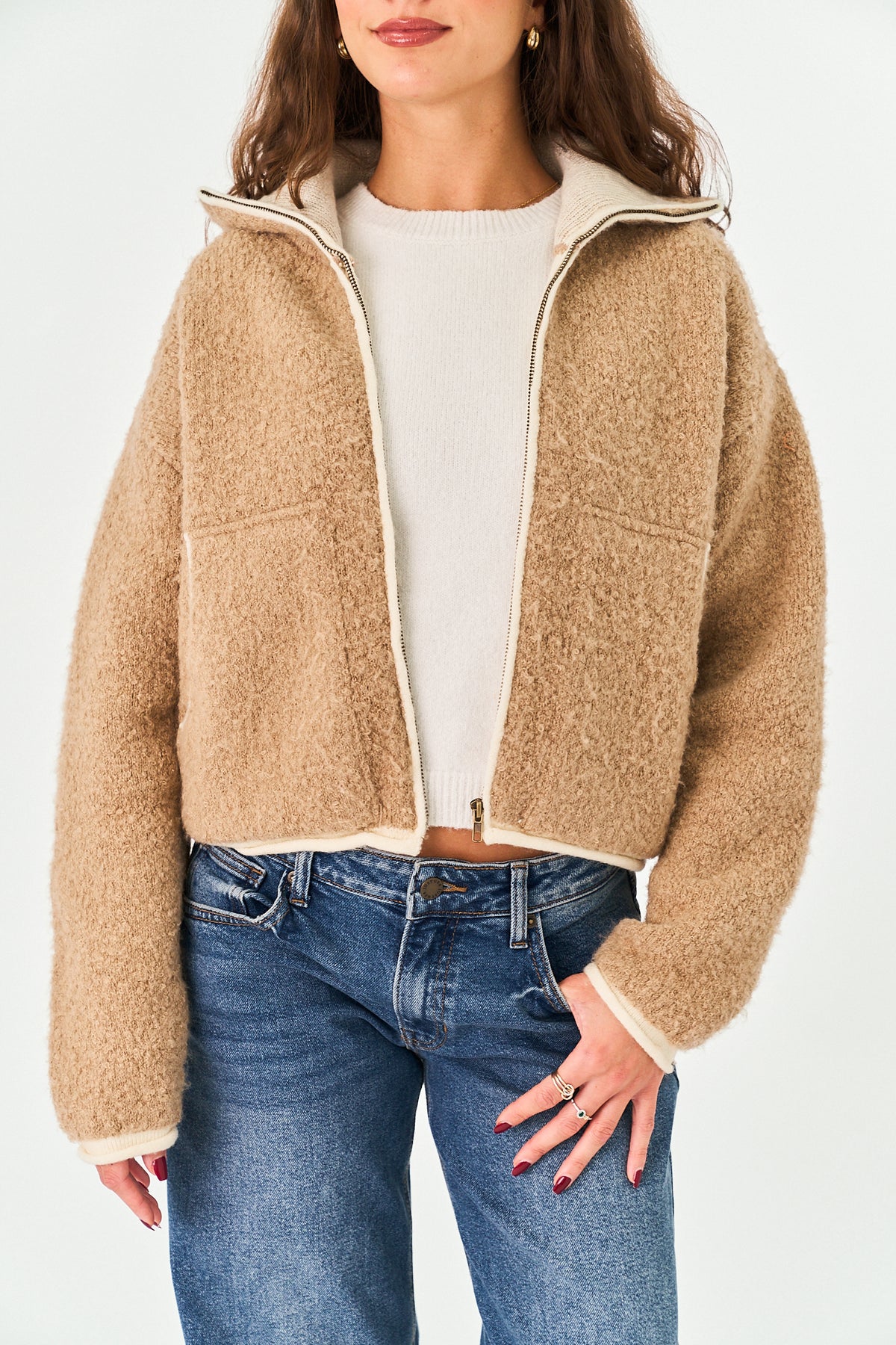 A woman wearing a cropped, fuzzy sherpa-style zip-up jacket in camel beige with cream trim, layered over a cream top and paired with blue jeans, against a plain light background.