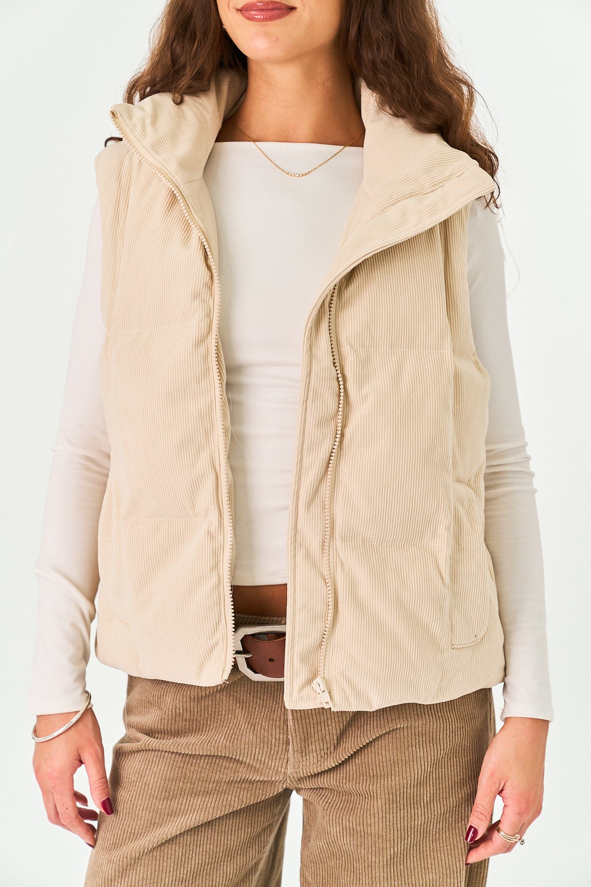 A woman wearing a light beige quilted puffer vest over a white long-sleeve top and brown corduroy pants, shown from the nose down against a plain white background.