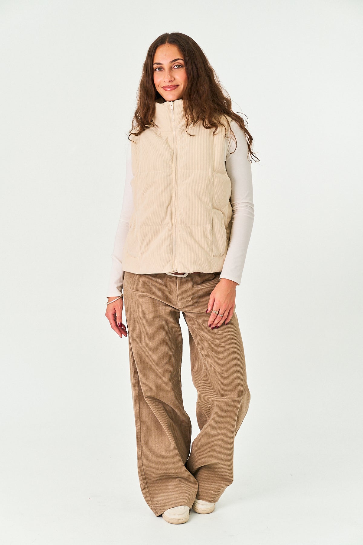 A woman modeling a beige zippered puffer vest over a white long-sleeve top and brown wide-leg corduroy pants, standing against a plain white studio background.
