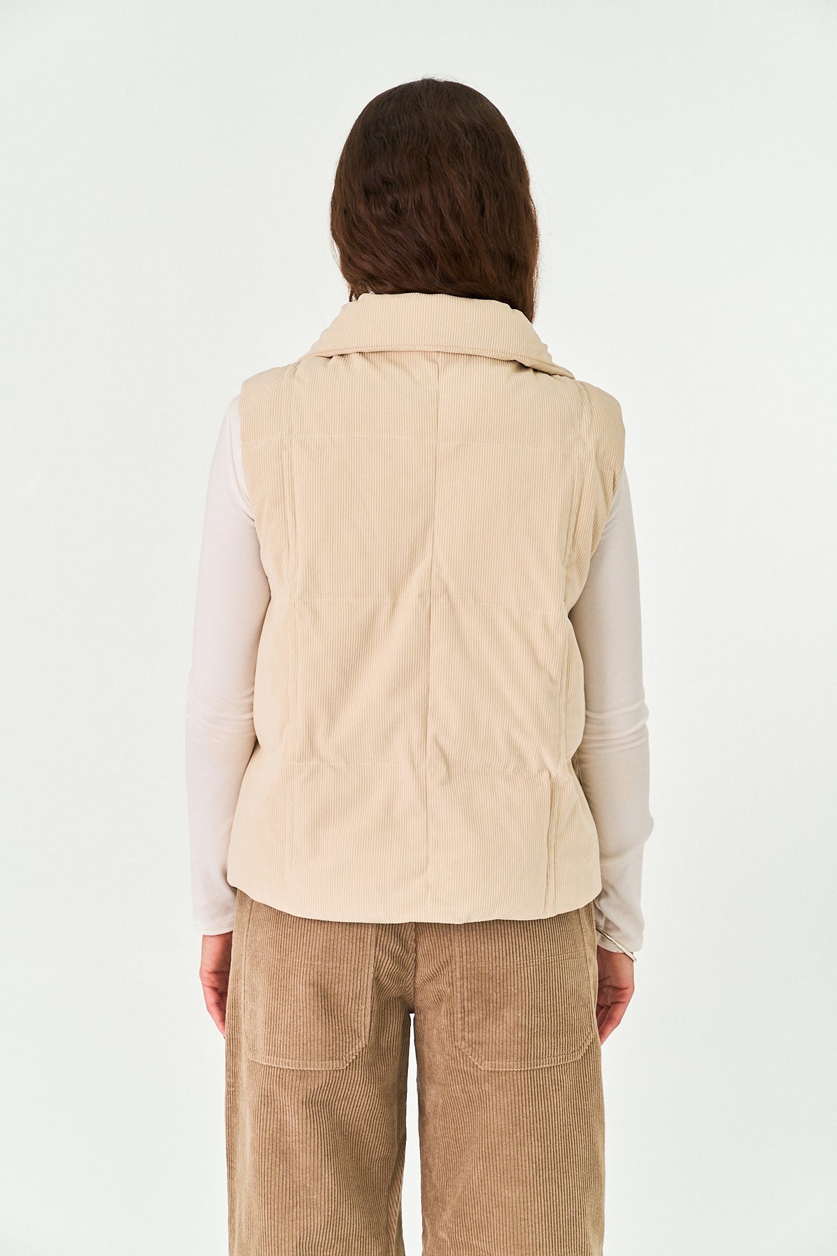 A person shown from the back wearing a light beige quilted puffer vest over a white long-sleeve top and tan corduroy pants, against a plain white studio background.