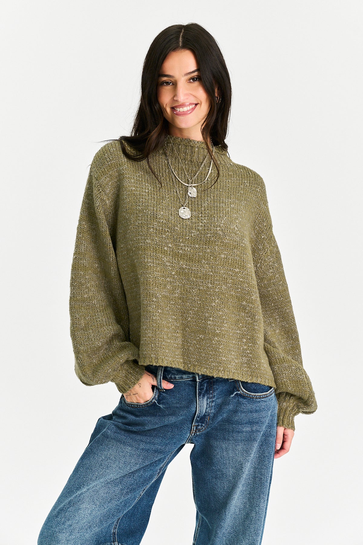A woman wearing a textured olive green mock-neck knit sweater with balloon sleeves and blue jeans, standing against a light gray studio background.