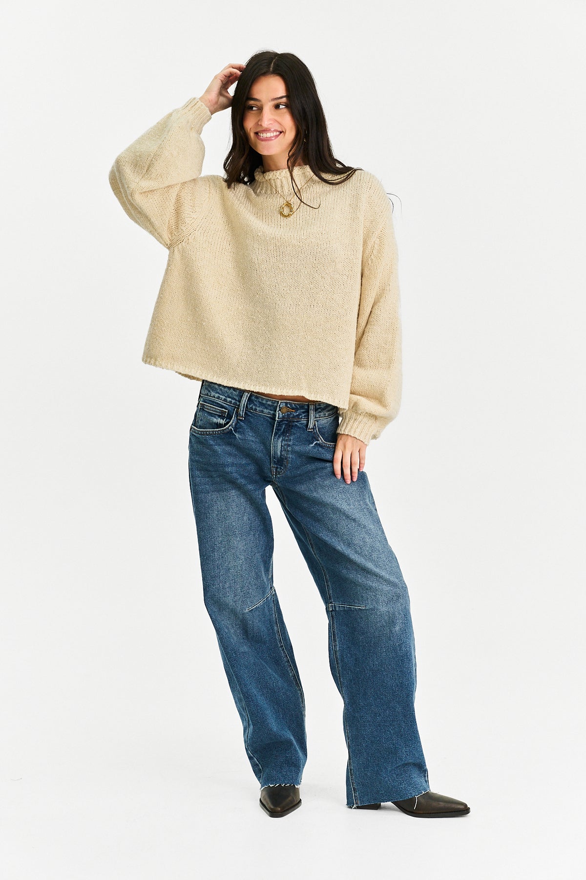 A woman modeling a cropped, long-sleeve knit sweater in a light beige or cream color, paired with blue wide-leg jeans and dark brown shoes, against a plain white studio background.