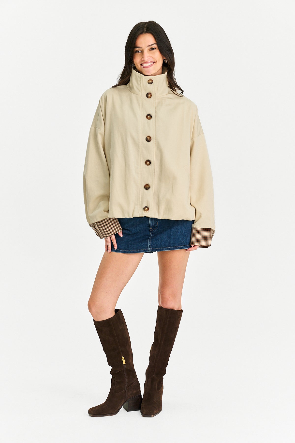 A woman modeling a beige high-neck button-front jacket with brown knit cuffs, paired with a blue denim mini skirt and dark brown knee-high boots, against a plain white studio background.