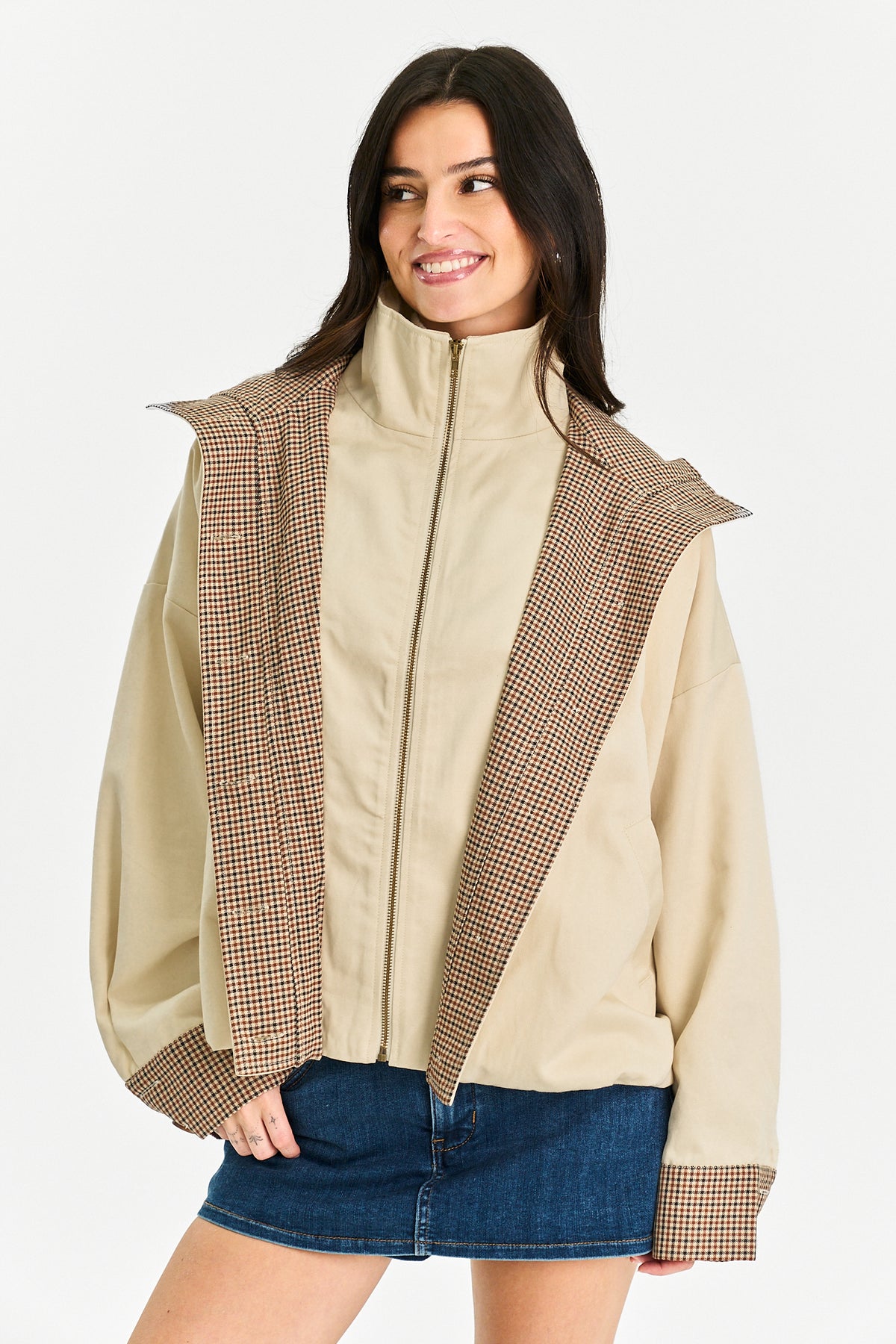 A woman wearing a beige zip-up jacket with brown and beige checkered patterned panels on the shoulders and cuffs, paired with a blue denim mini skirt, against a plain light gray background.