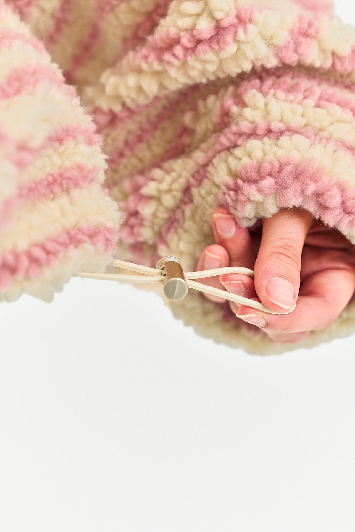 A close-up of a person's hand adjusting a drawstring toggle on a chunky, fuzzy fleece jacket in pink and cream stripes, set against a solid white background.