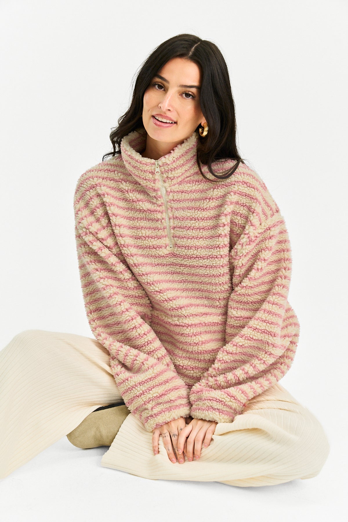 A woman sits cross-legged on a white studio background wearing a fuzzy quarter-zip pullover sweater with horizontal pink and beige stripes, paired with cream ribbed pants.