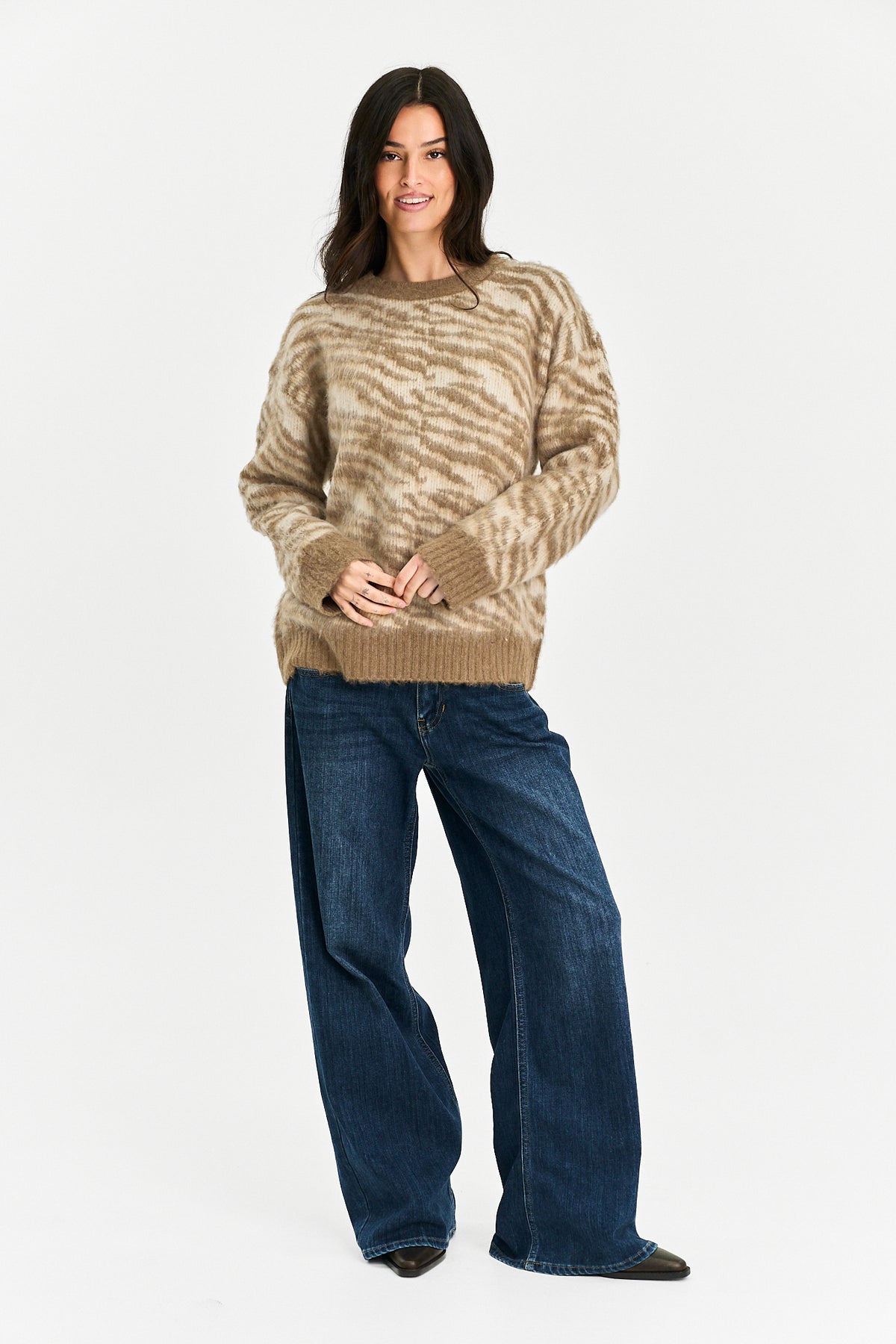 A woman modeling a beige and brown wavy-patterned knit sweater with wide-leg blue jeans, standing against a plain white studio background.