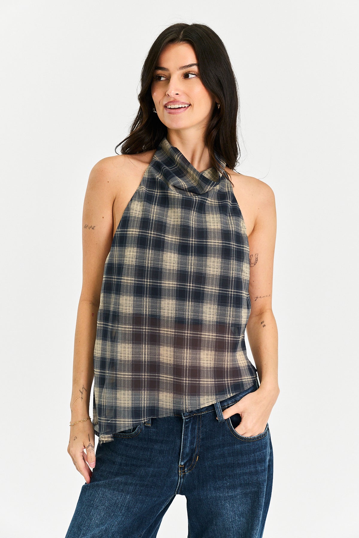 A woman modeling a sleeveless halter-neck plaid top in navy, beige, and brown tones, paired with dark blue jeans, against a plain light gray background.
