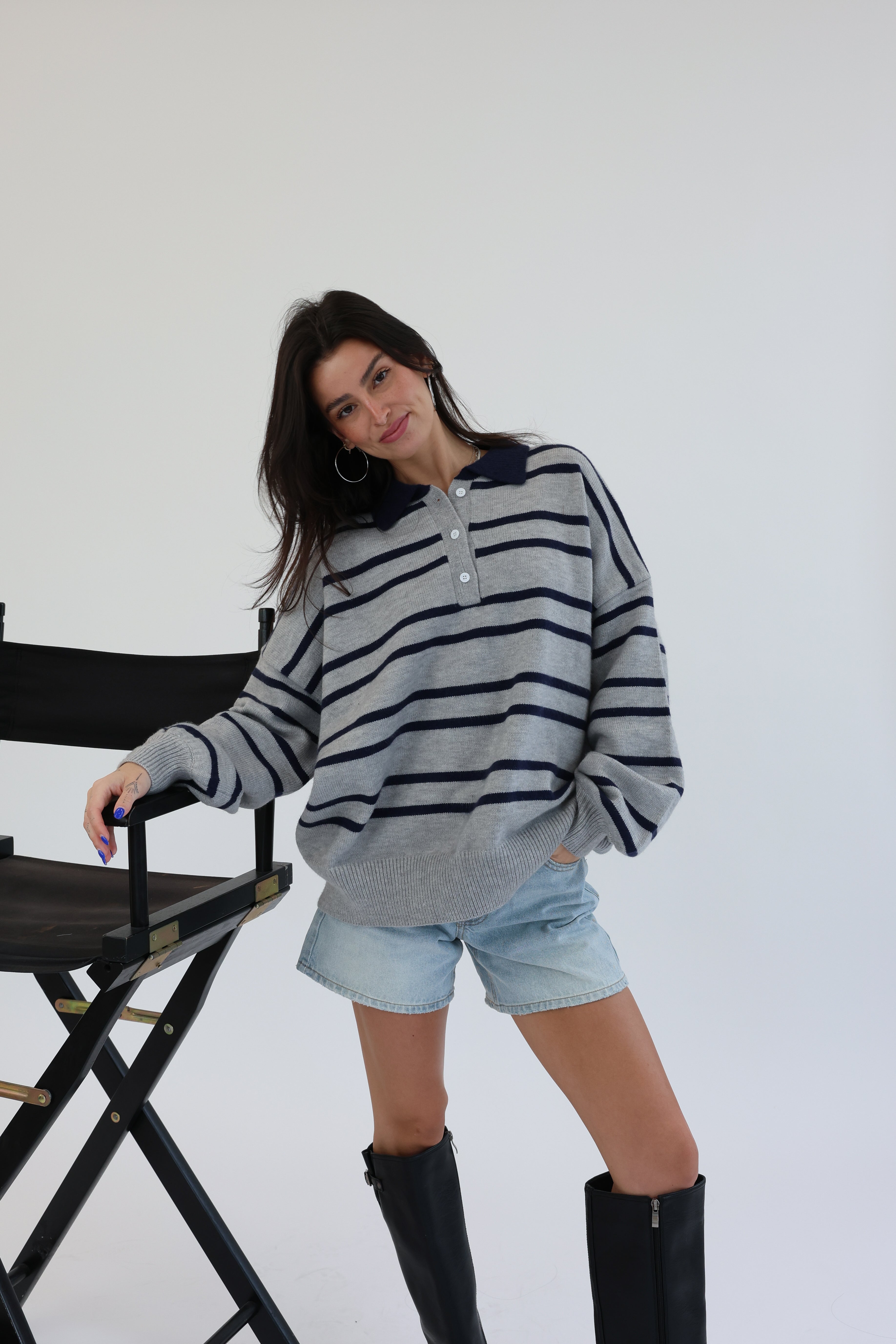 Woman wearing a striped sweater and denim shorts standing next to a chair on a white background