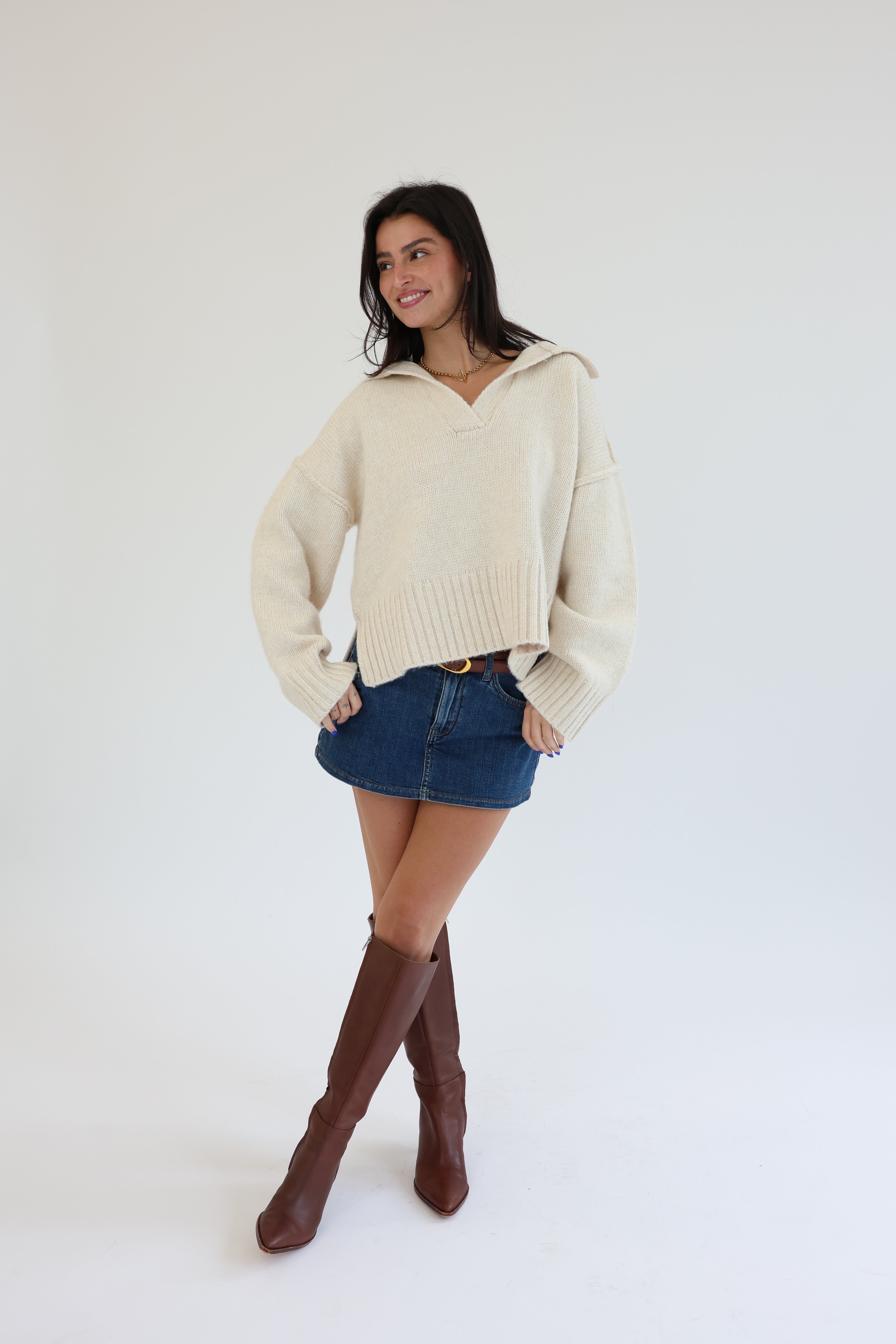 Woman wearing a cream sweater, denim skirt, and brown knee-high boots on a white background