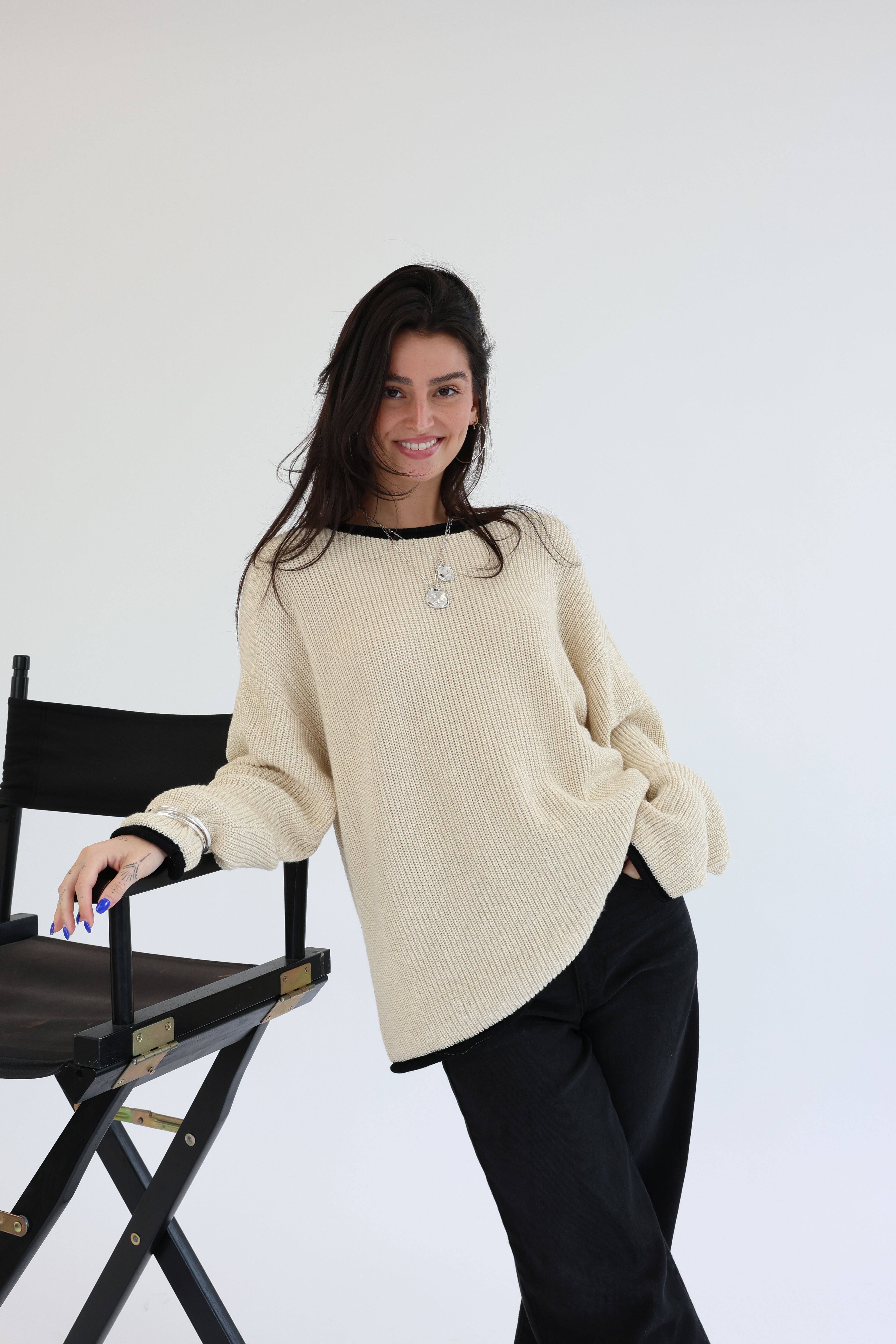 Woman wearing a beige sweater and black pants standing next to a director's chair on a white background