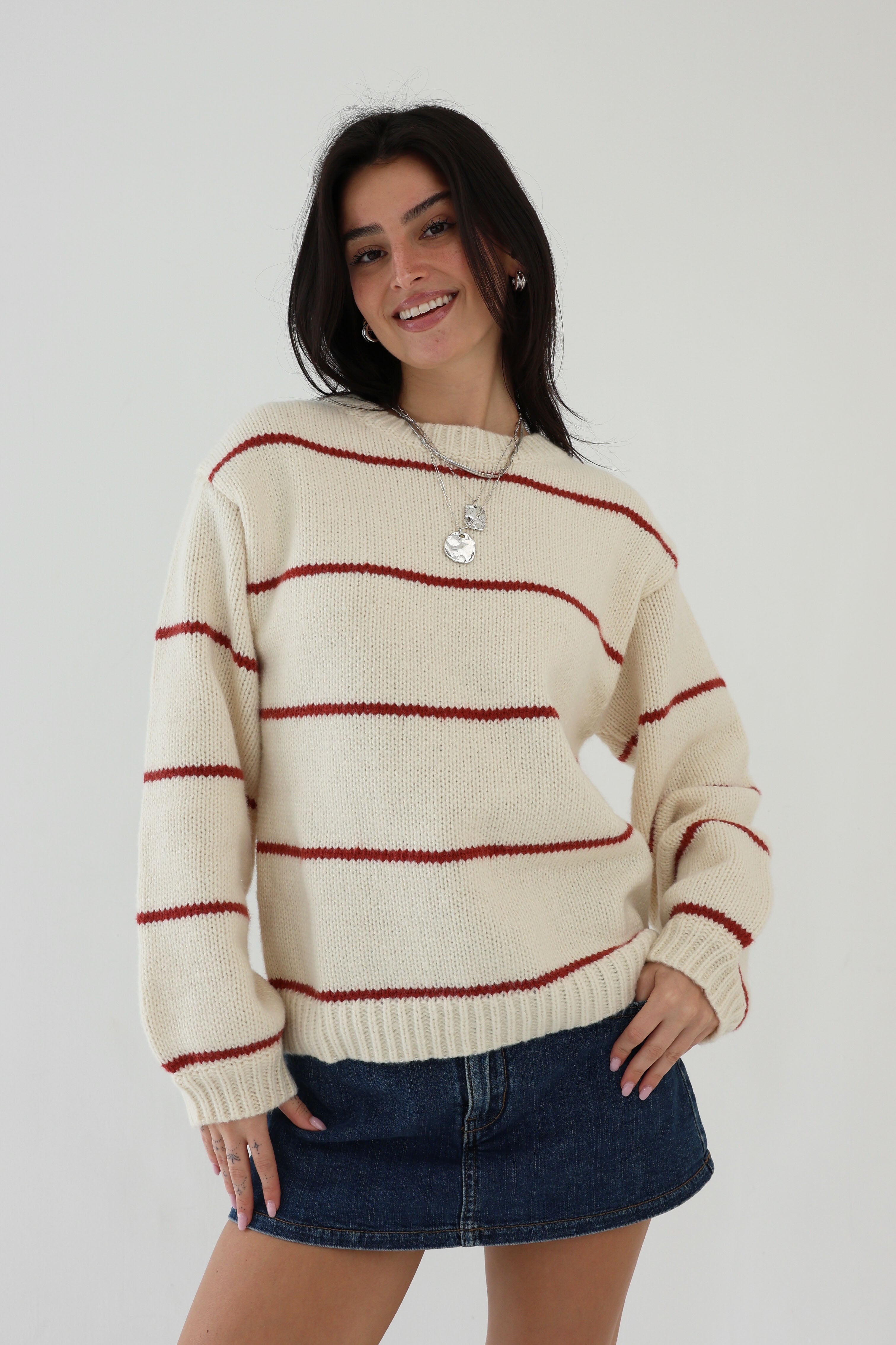 Woman wearing a cream sweater with red stripes and a denim skirt against a plain background