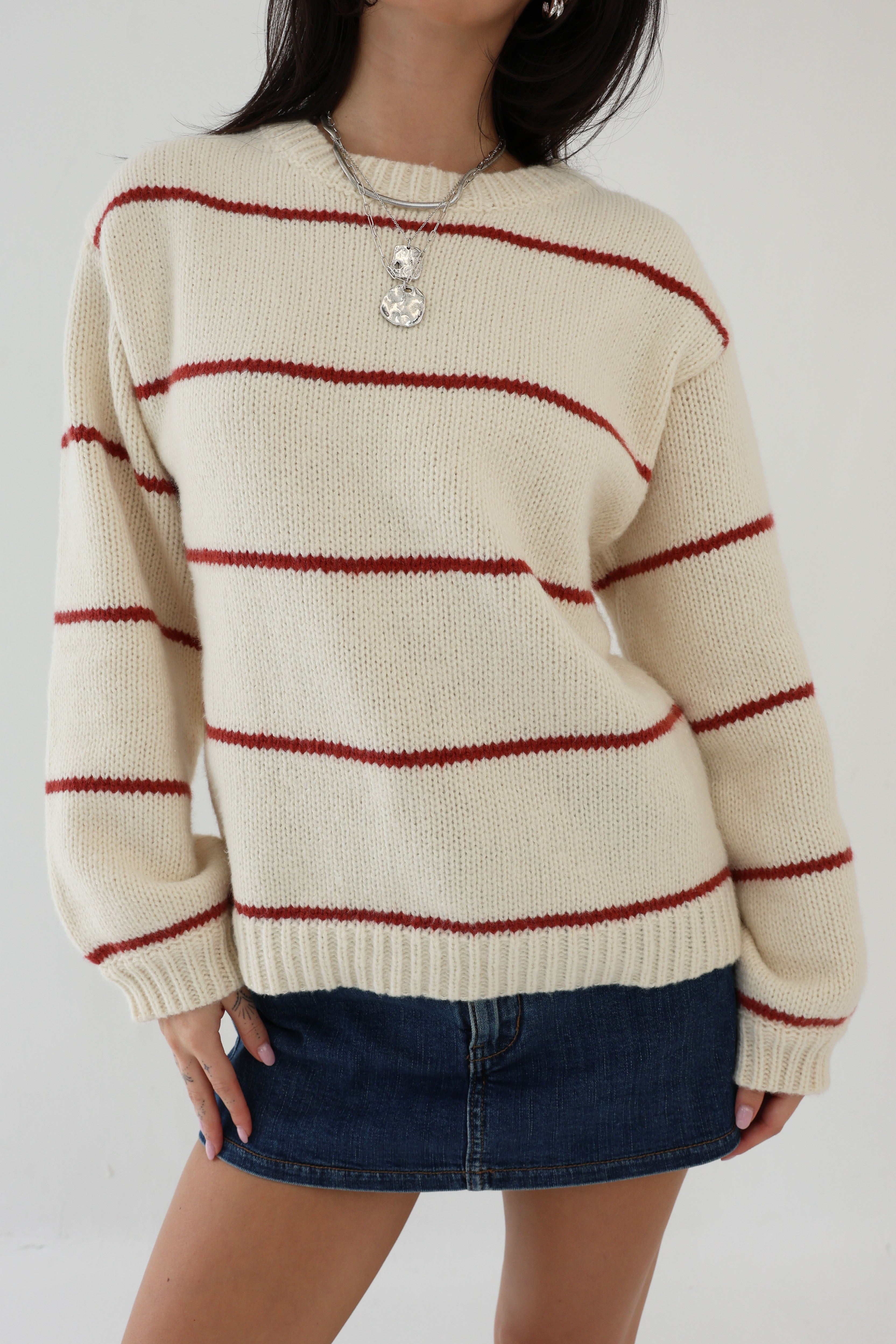 Person wearing a cream sweater with red stripes and a denim skirt on a plain background