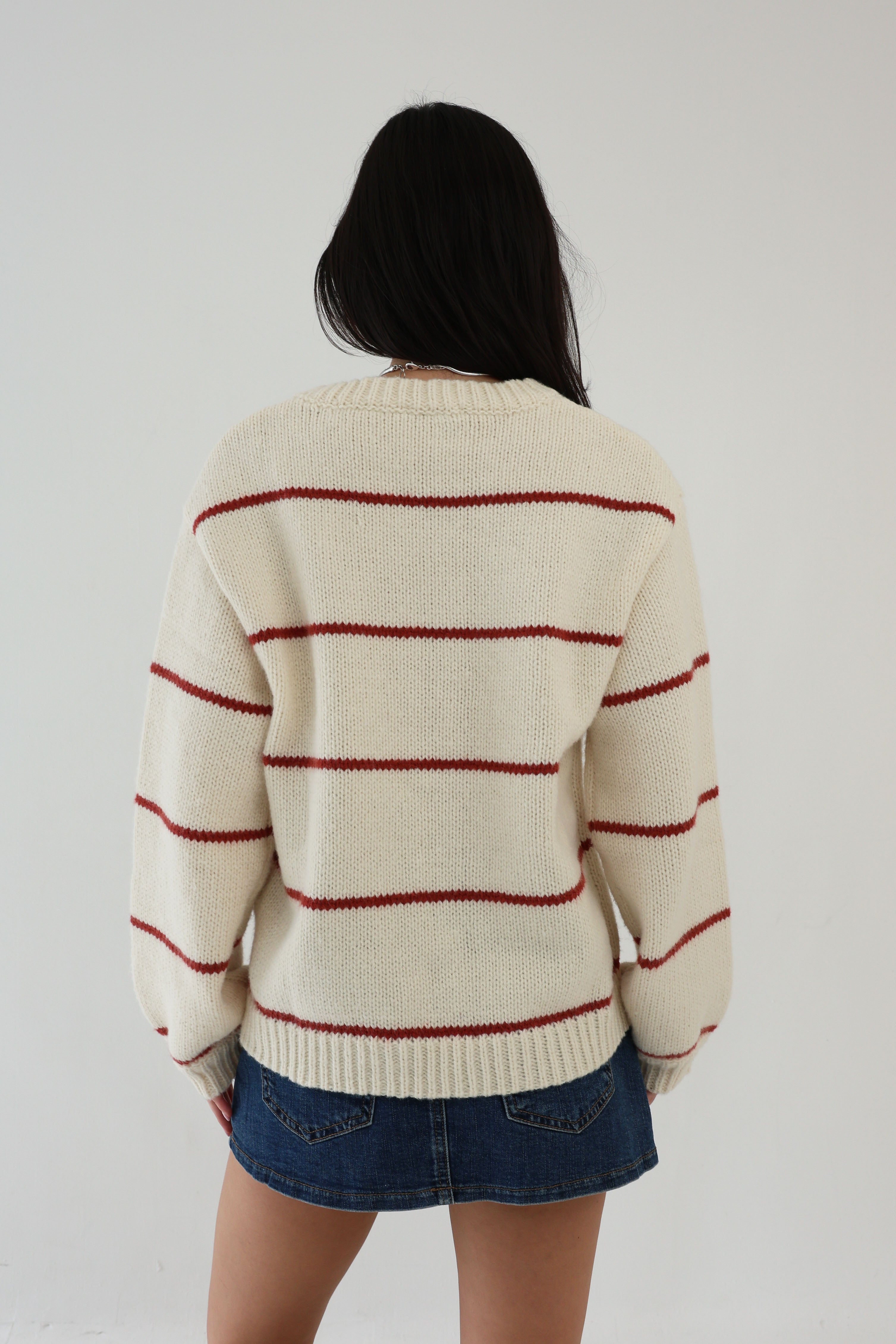 Person wearing a cream sweater with red stripes and a denim skirt on a plain background