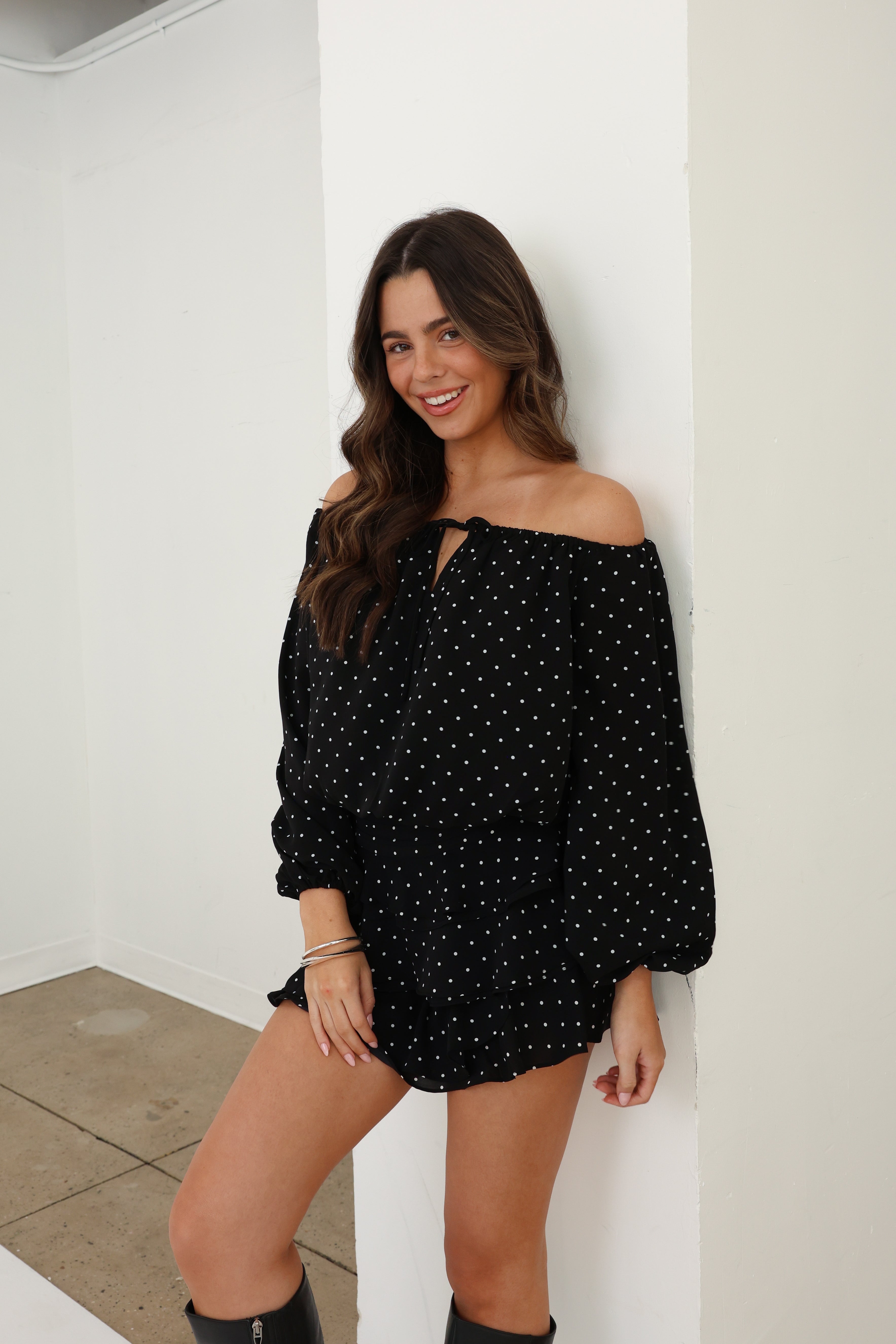Woman wearing a black off-shoulder dress with white polka dots against a white wall.
