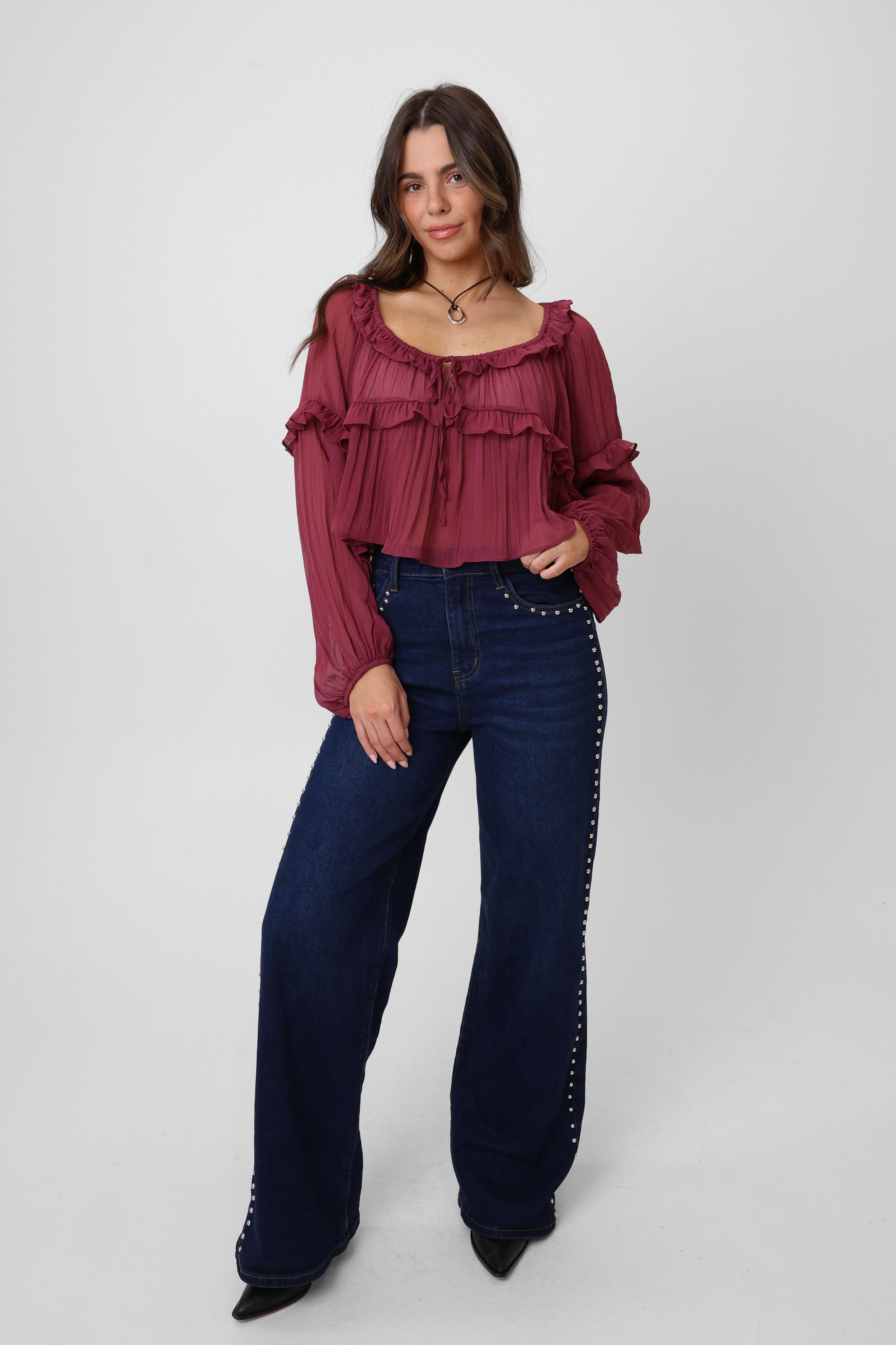 Woman wearing a burgundy ruffled blouse and dark blue jeans with studded details on a white background