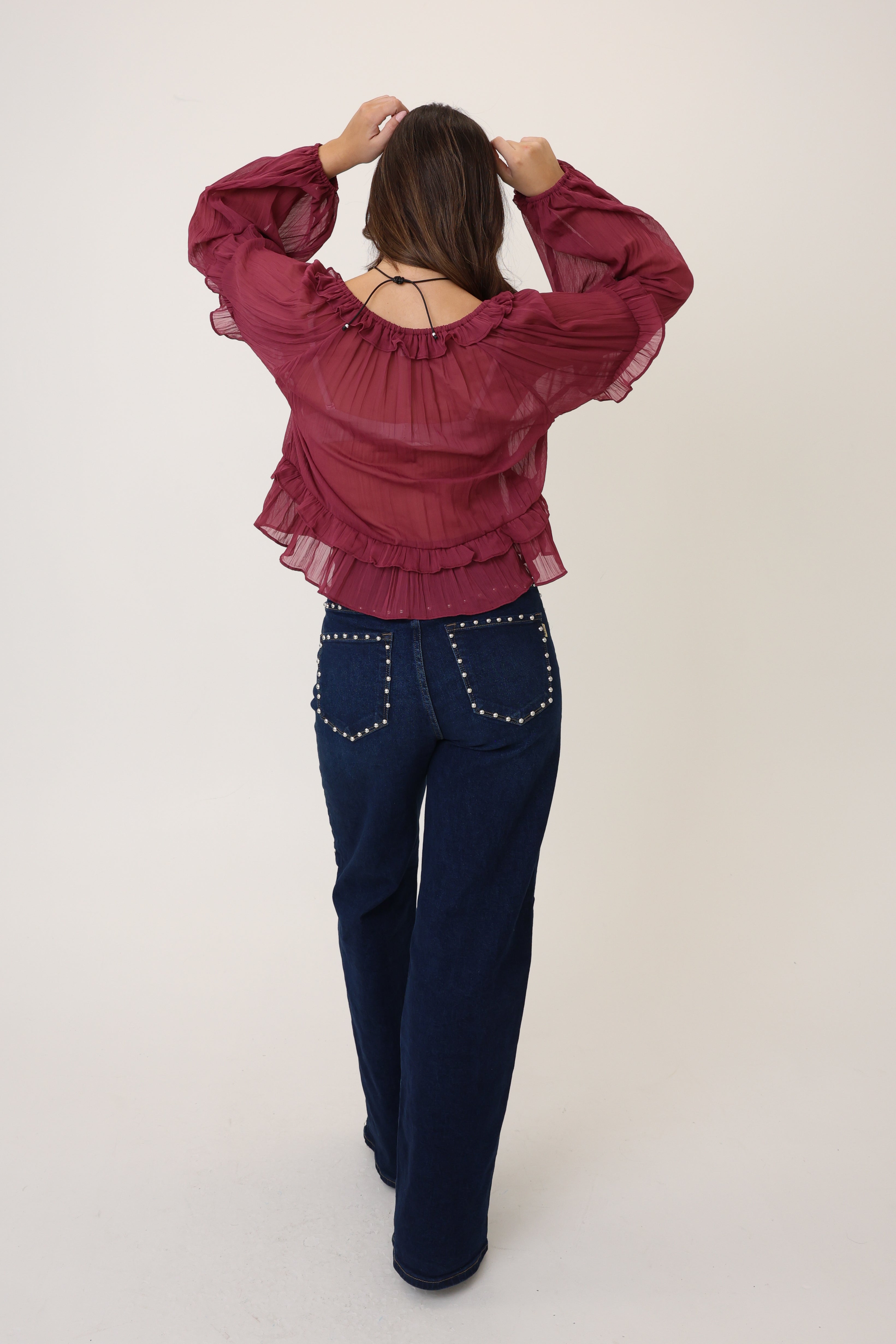 Person wearing a burgundy blouse with ruffled details and dark blue pants on a white background