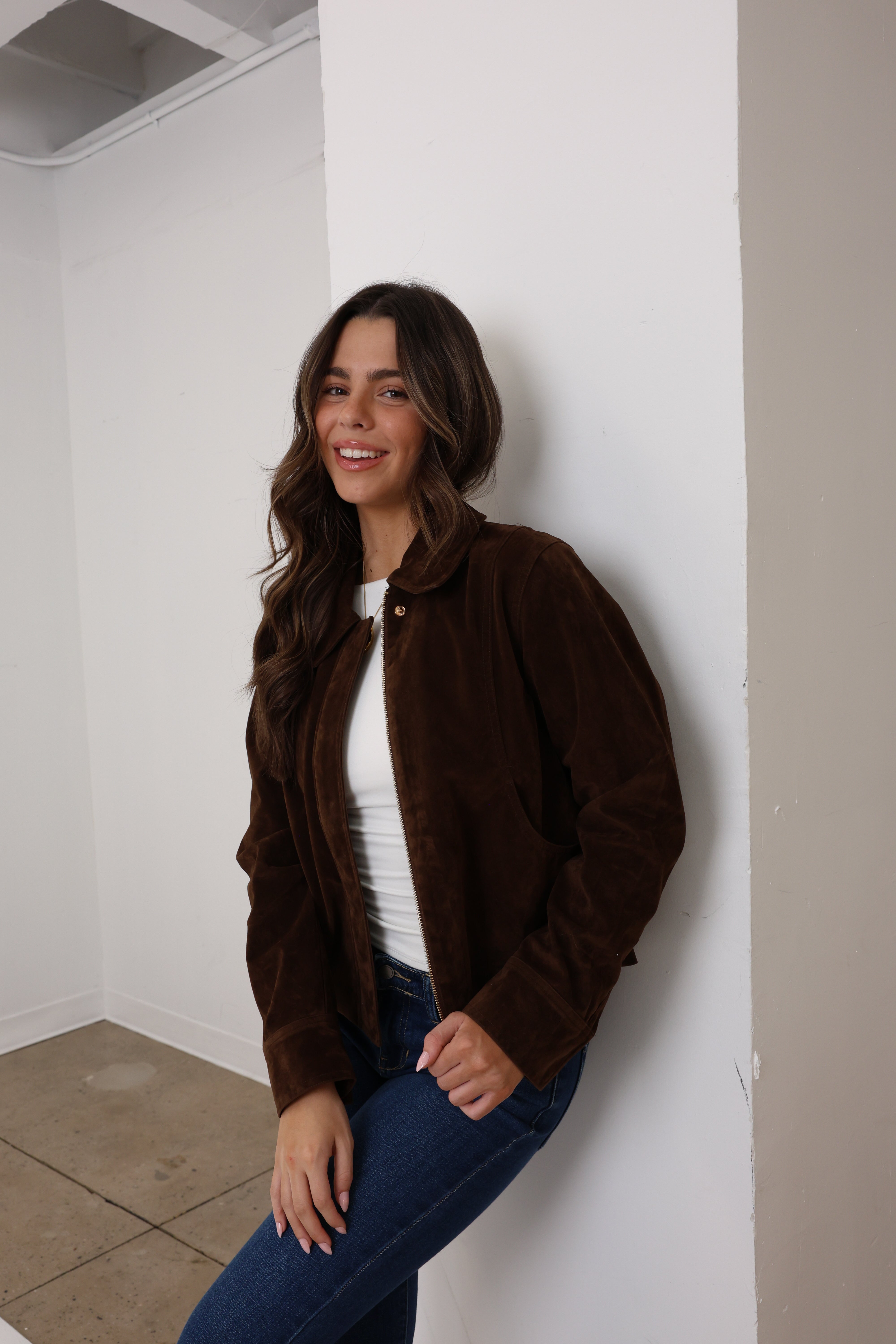 Woman wearing a brown jacket and blue jeans leaning against a white wall.