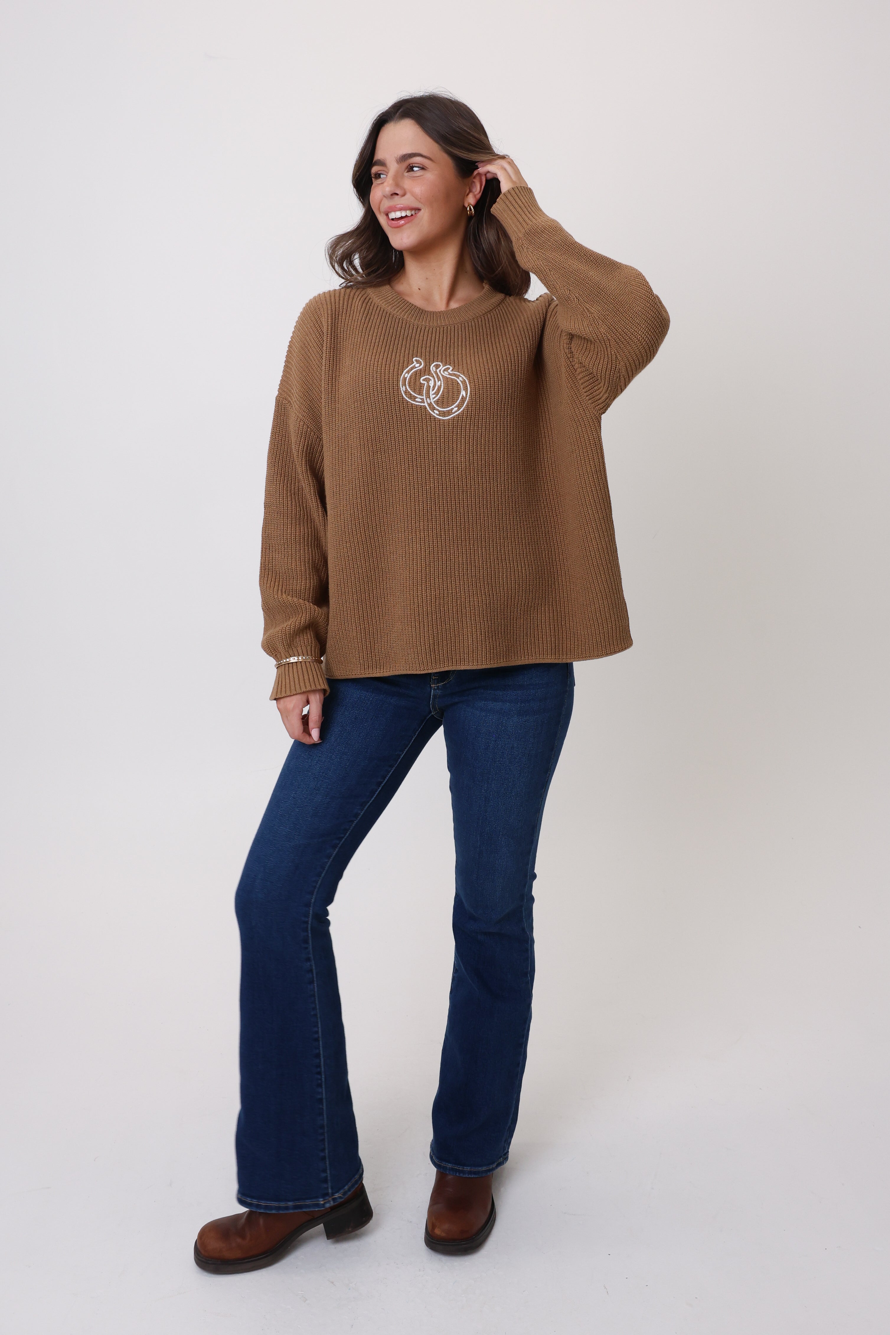 Woman wearing a brown sweater with a logo and blue jeans on a white background