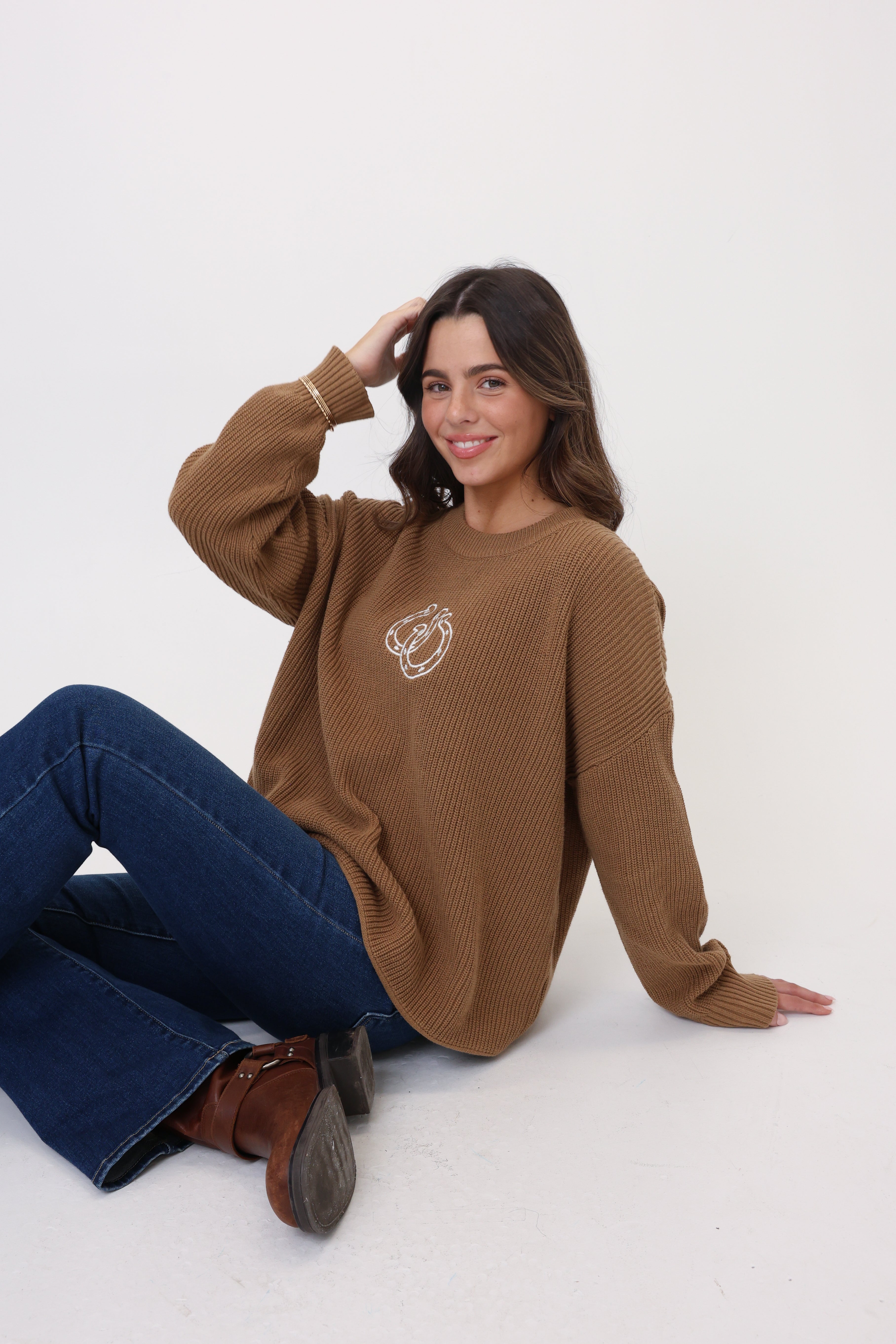 Woman wearing a brown sweater with a logo, sitting on a white floor.