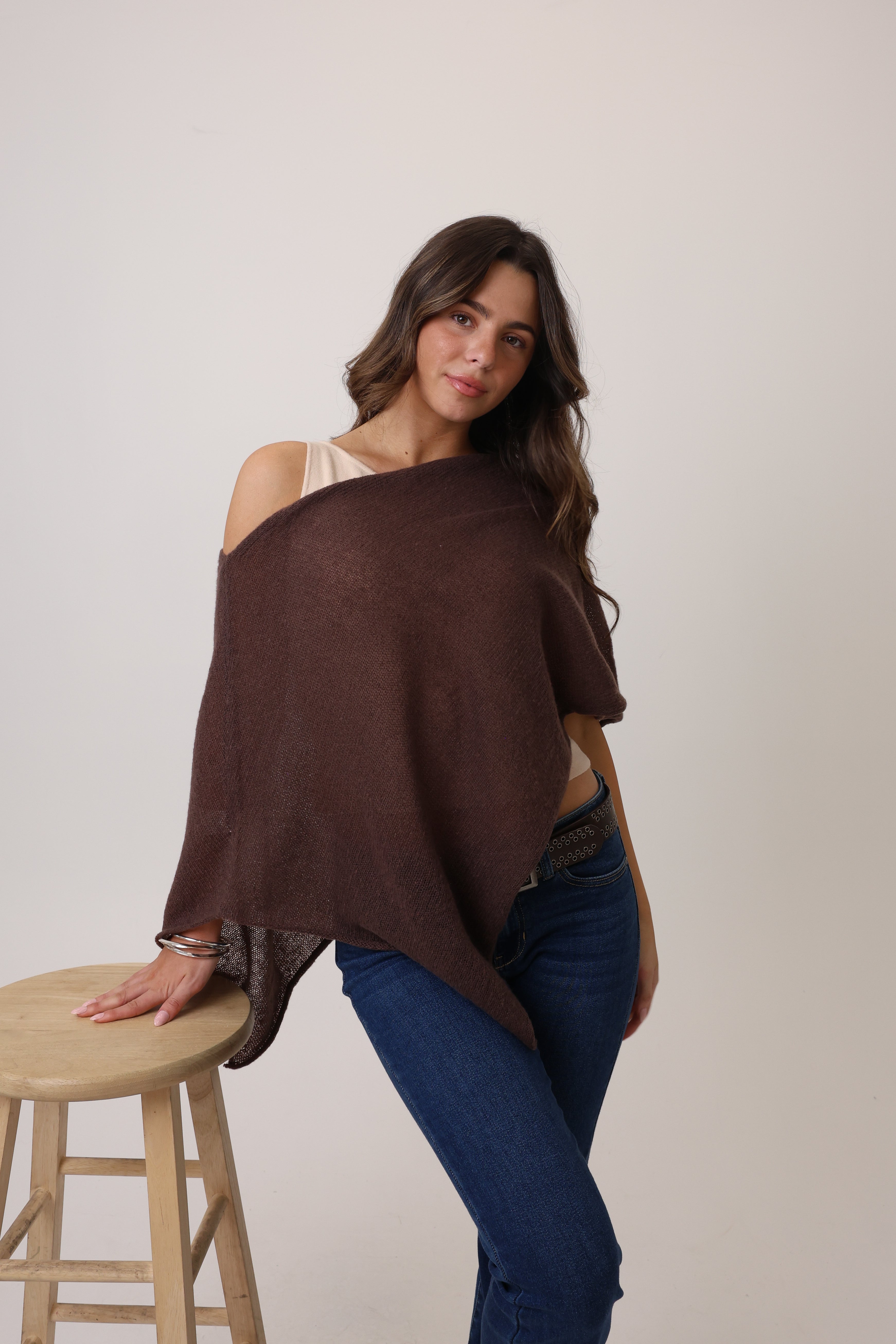 Woman wearing a brown poncho over one shoulder, standing next to a wooden stool against a plain background.