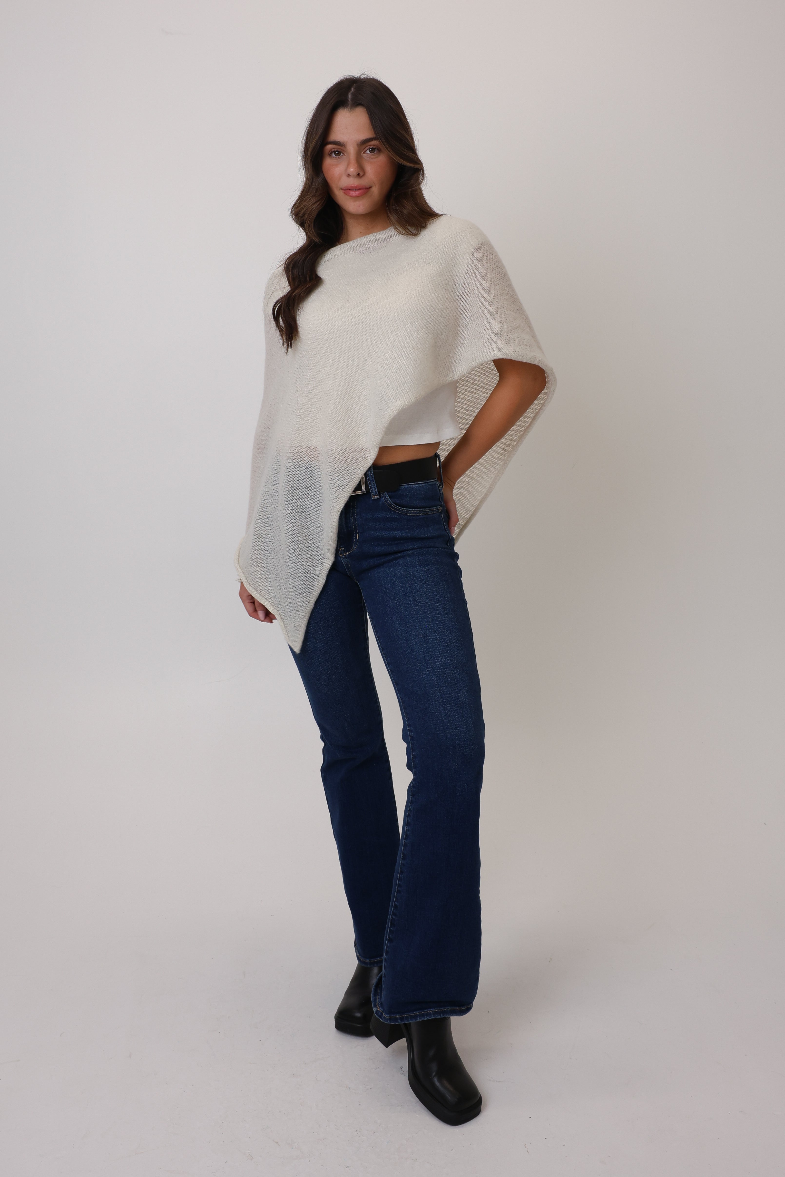 Woman wearing a white poncho, blue jeans, and black shoes on a white background