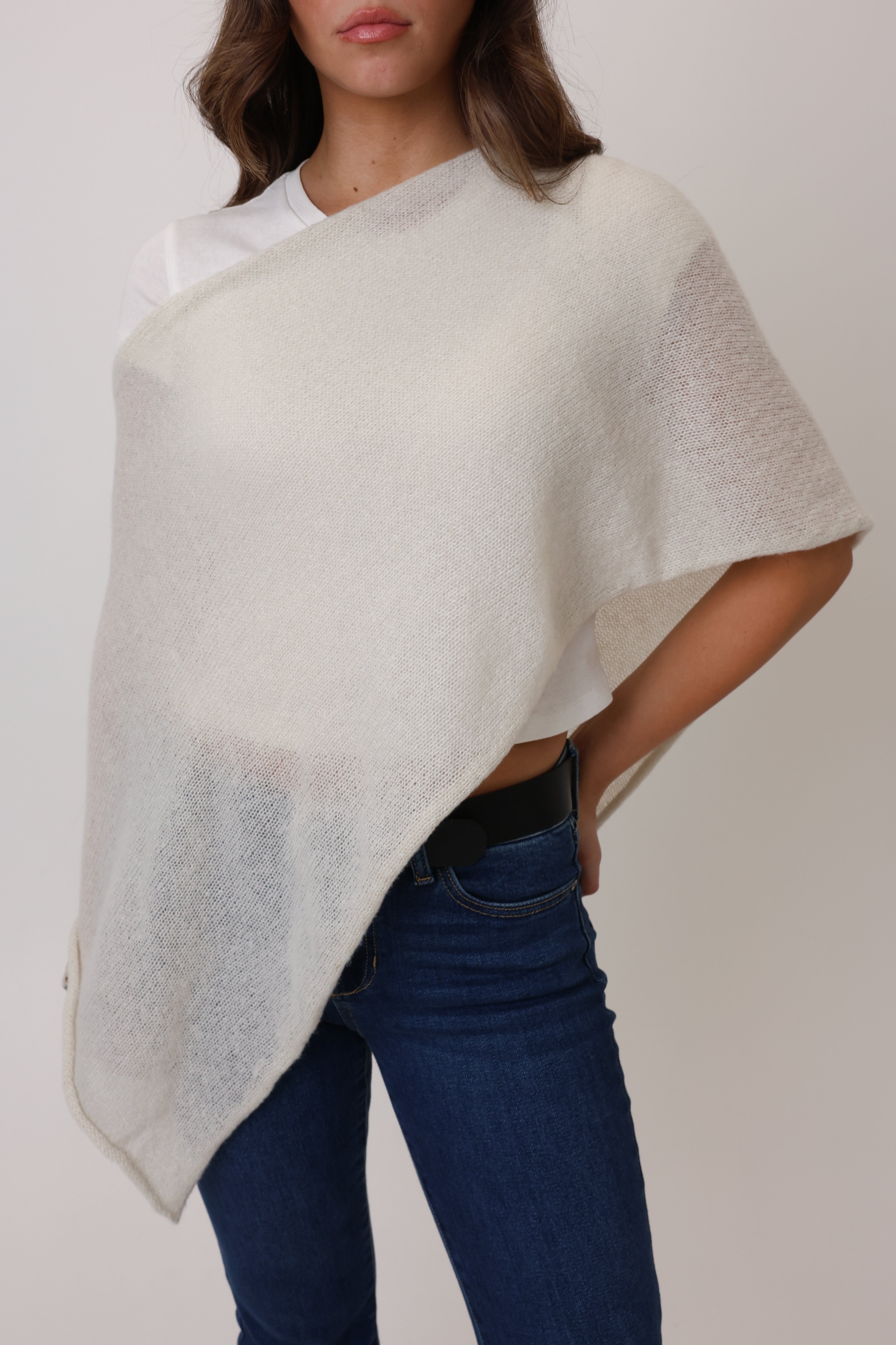 Woman wearing a light gray poncho over a white top and blue jeans on a plain background