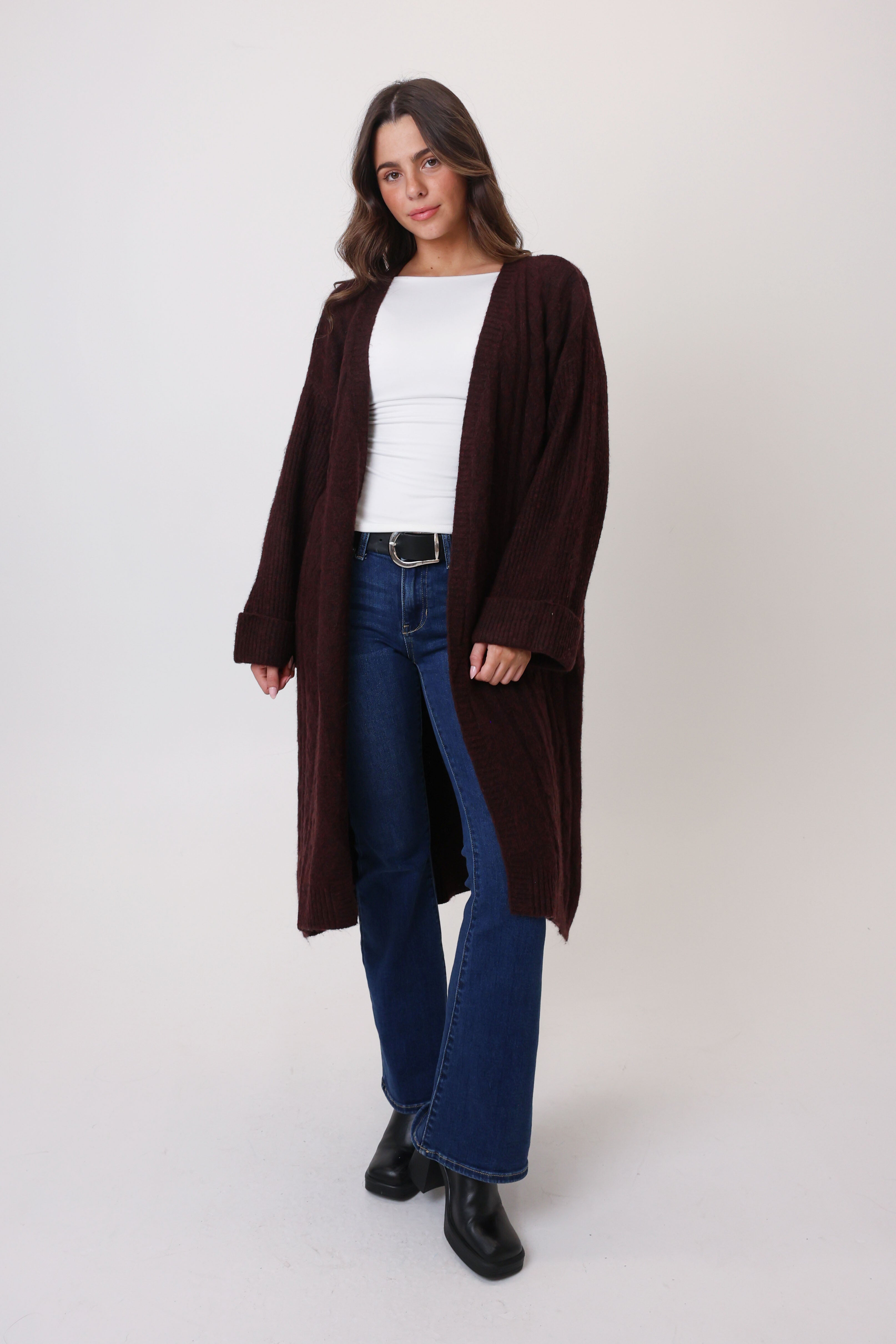 Woman wearing a long brown cardigan over a white top and blue jeans on a white background