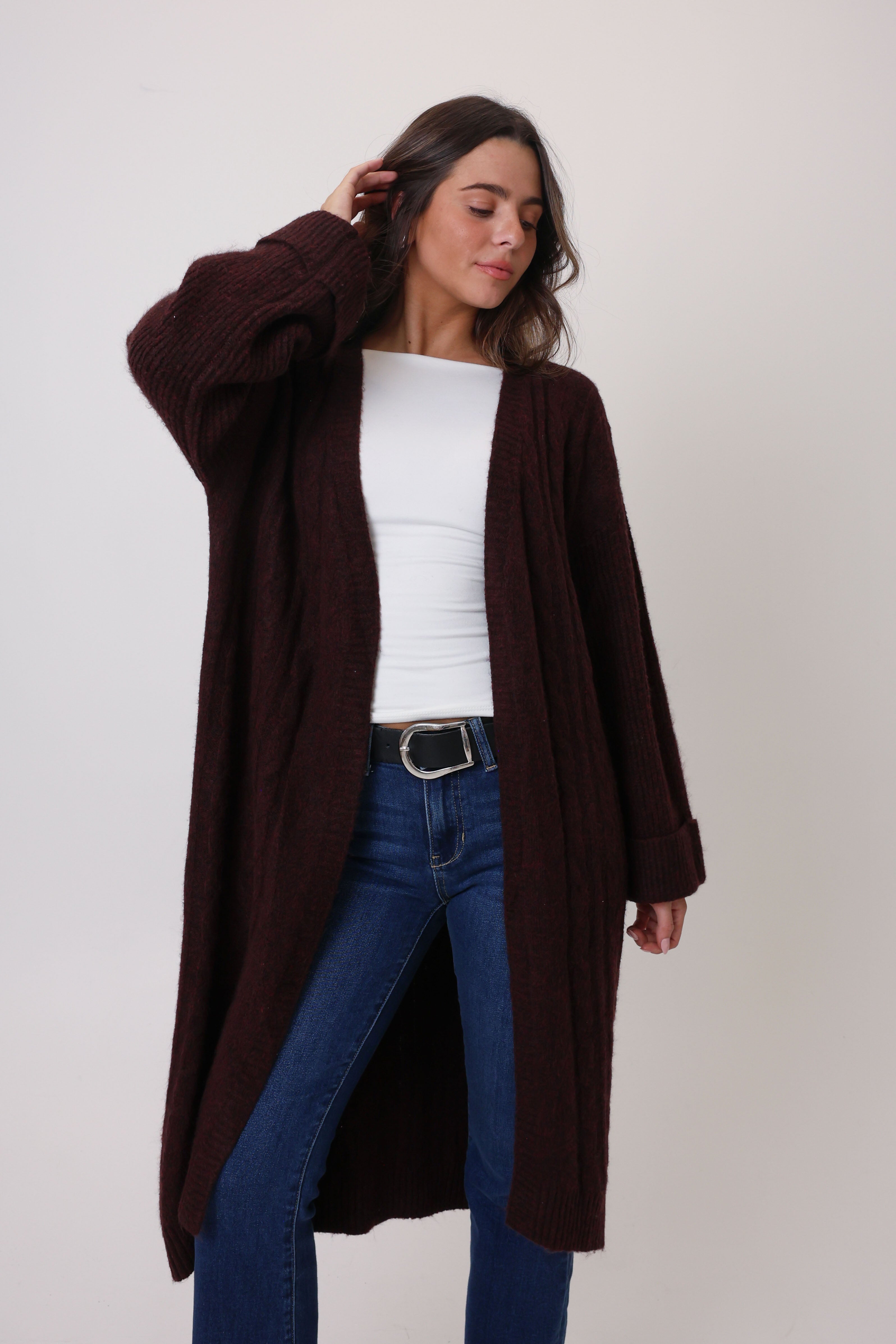 Woman wearing a brown cardigan over a white top and blue jeans on a plain background