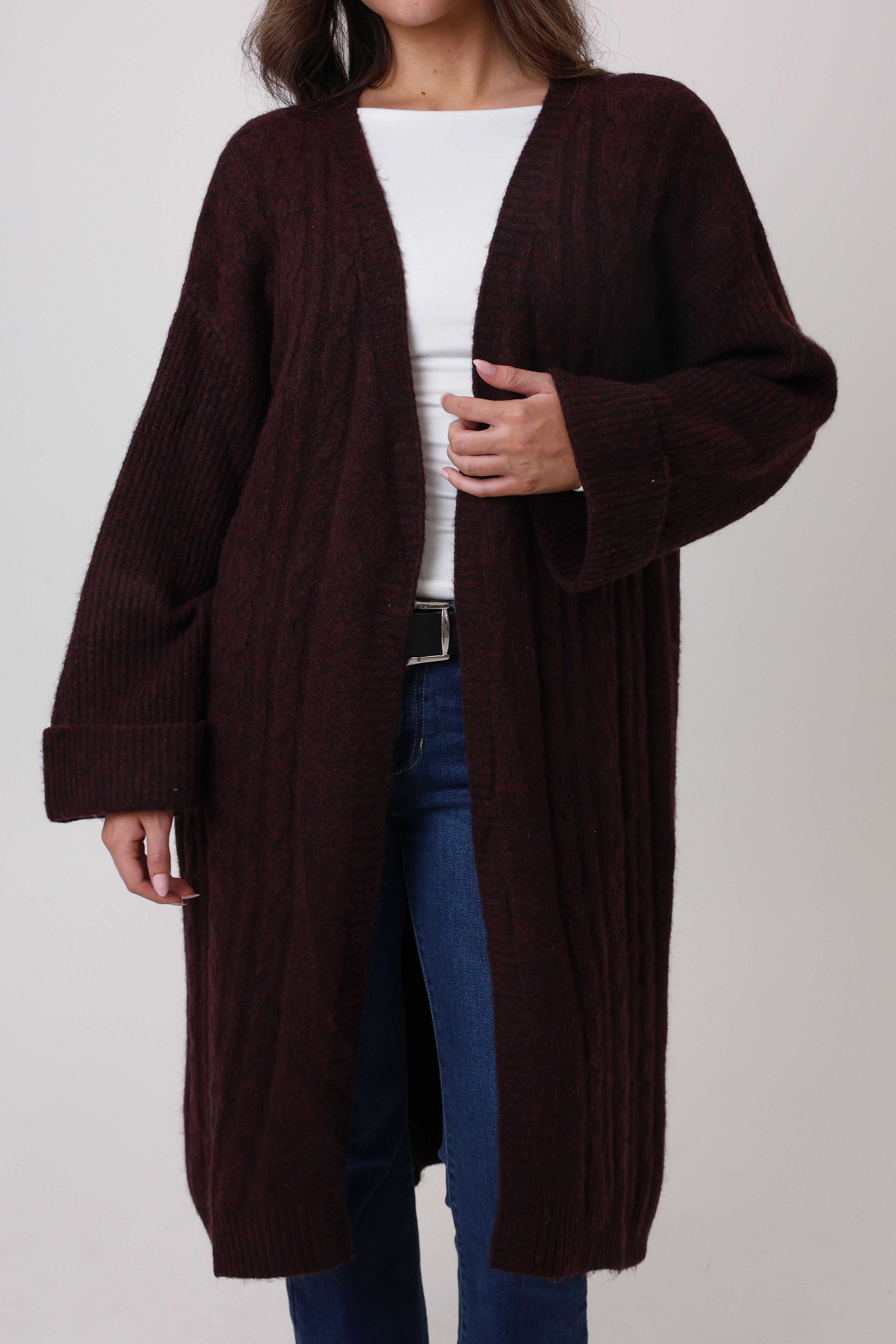 Person wearing a long brown cardigan over a white top and blue jeans on a plain background